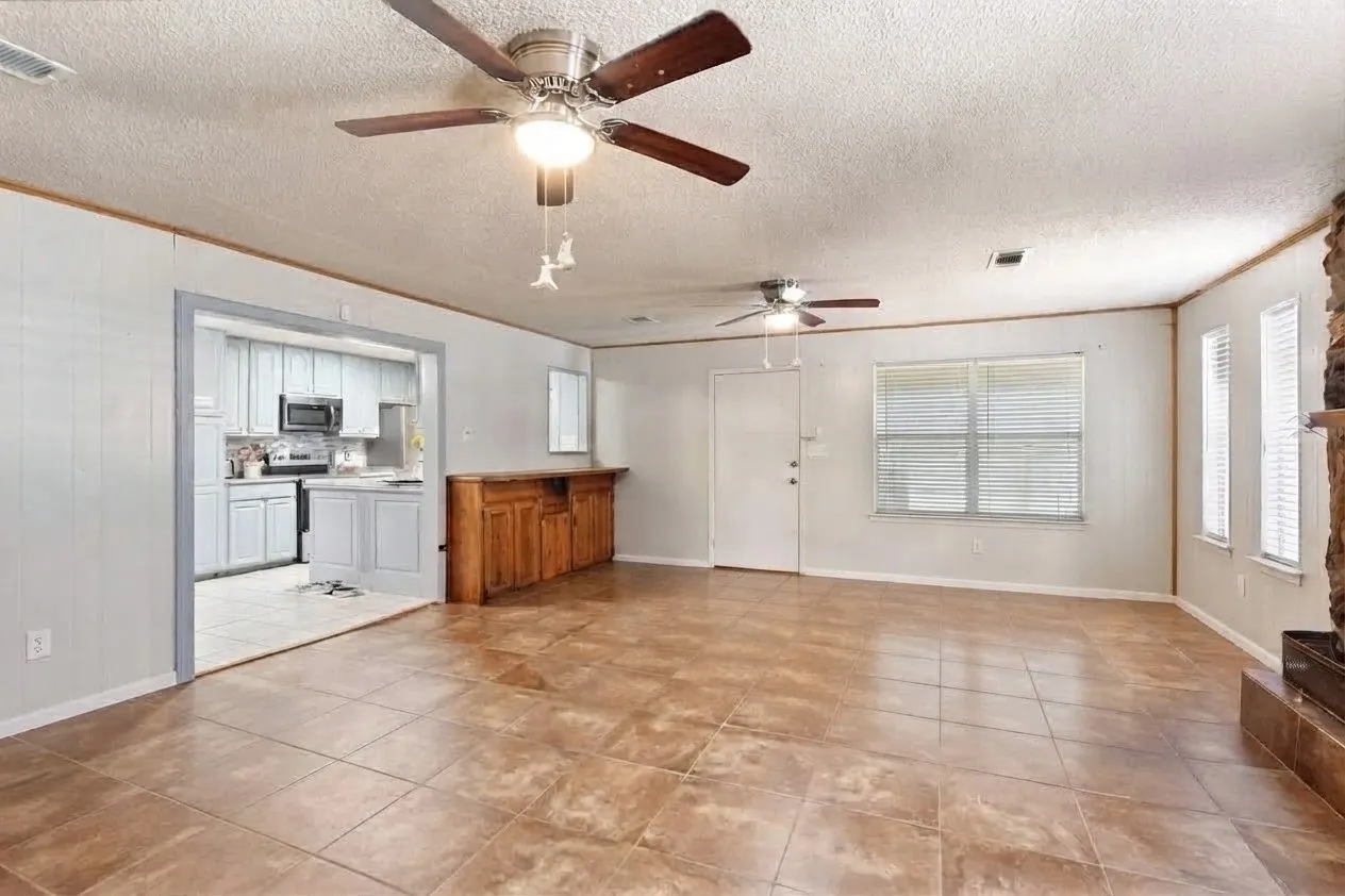 Spacious living area featuring tile flooring, two ceiling fans with integrated lighting, multiple windows with blinds, and a wood-finish cabinet