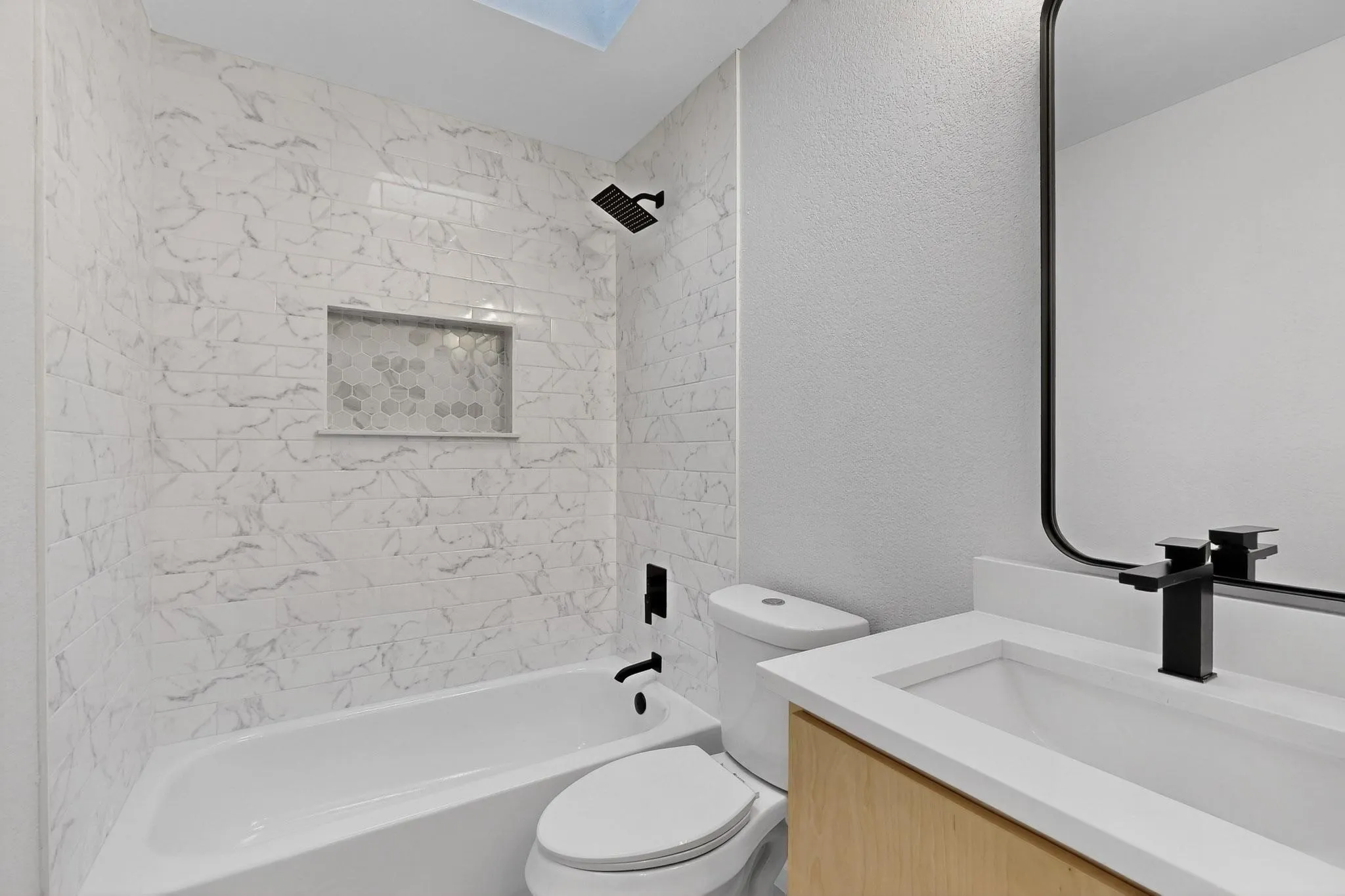White subway tile with subtle grey veining, shower niche with mosaic tile inlay, matte black fixtures, light wood-finish vanity, and a rectangular undermount sink