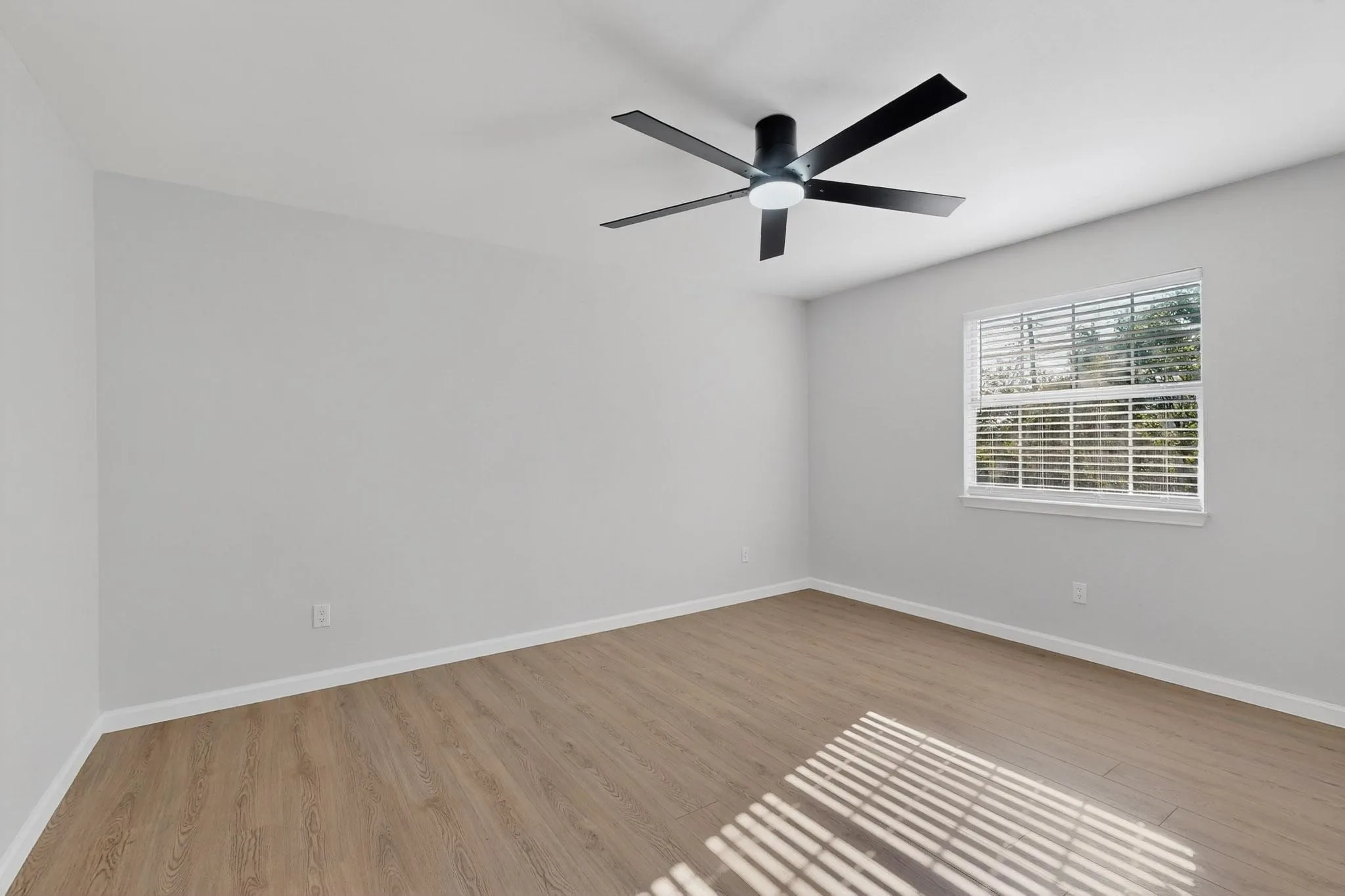 Spacious room featuring wood-finish flooring, light gray walls, a window with horizontal blinds, and a ceiling fan with integrated lighting