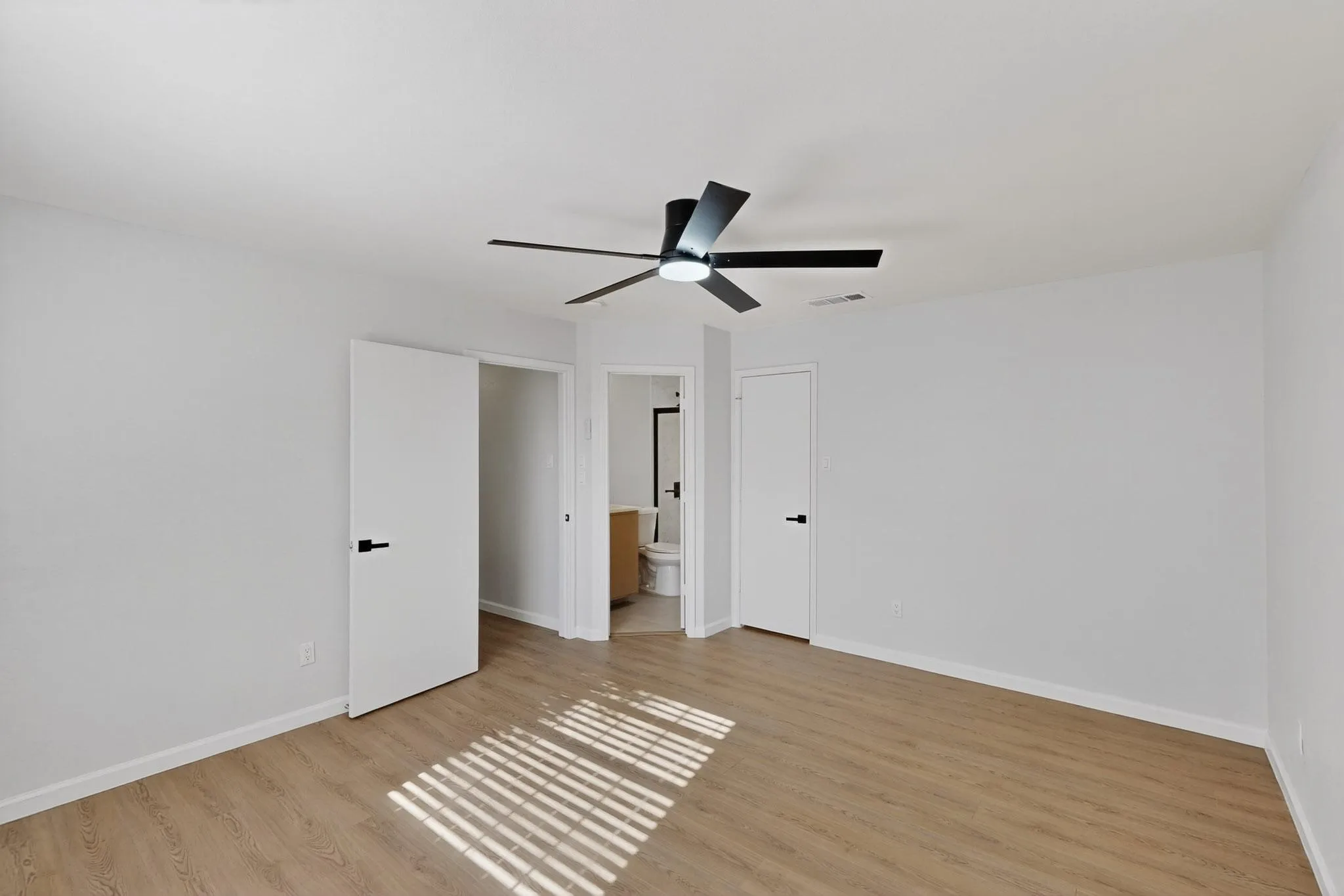 Expansive room featuring wood-finish flooring, a modern black ceiling fan with integrated lighting, and white walls