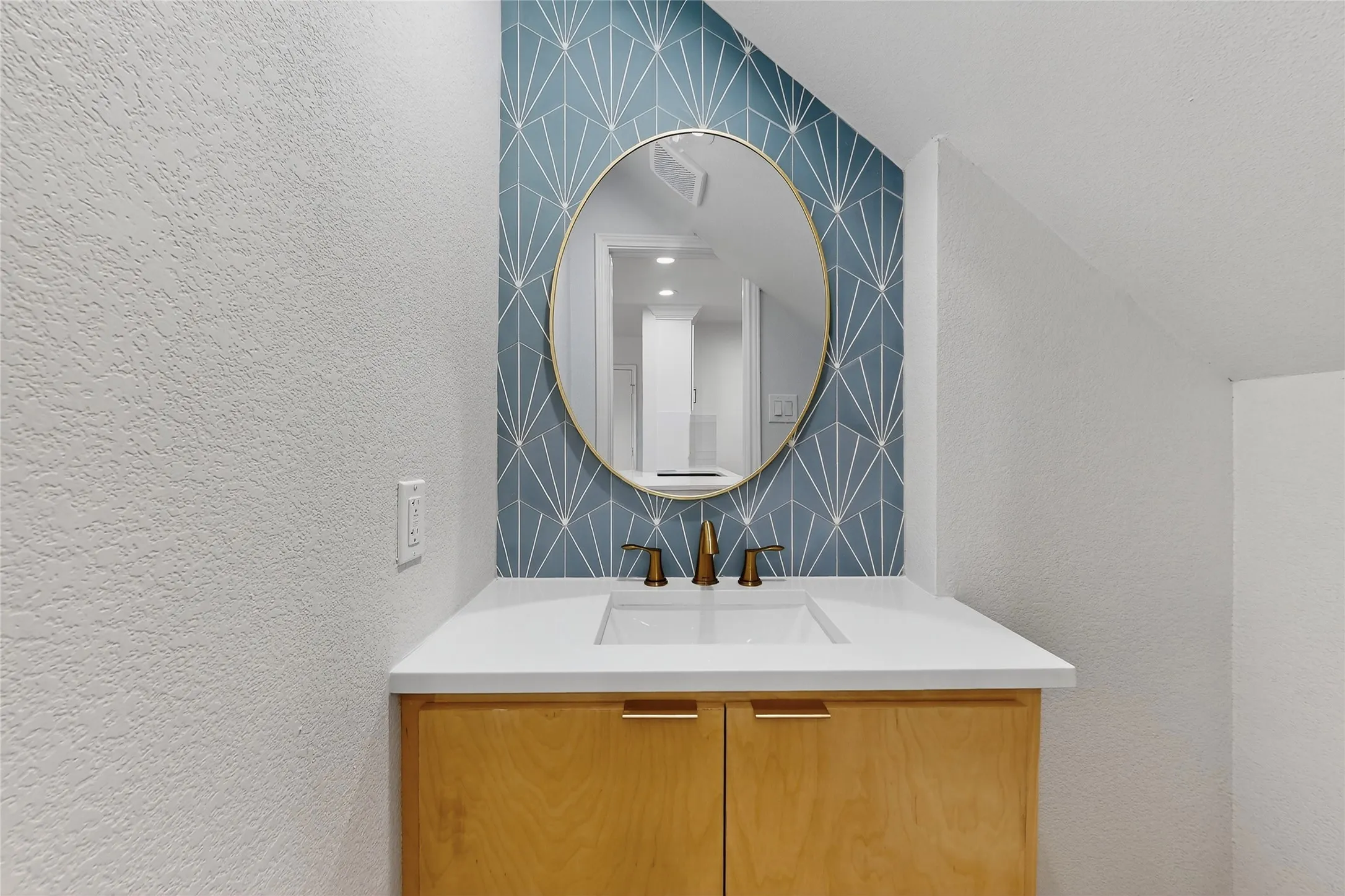 Recessed vanity with a built-in rectangular sink, light wood-finish cabinetry, and a gold-framed circular mirror