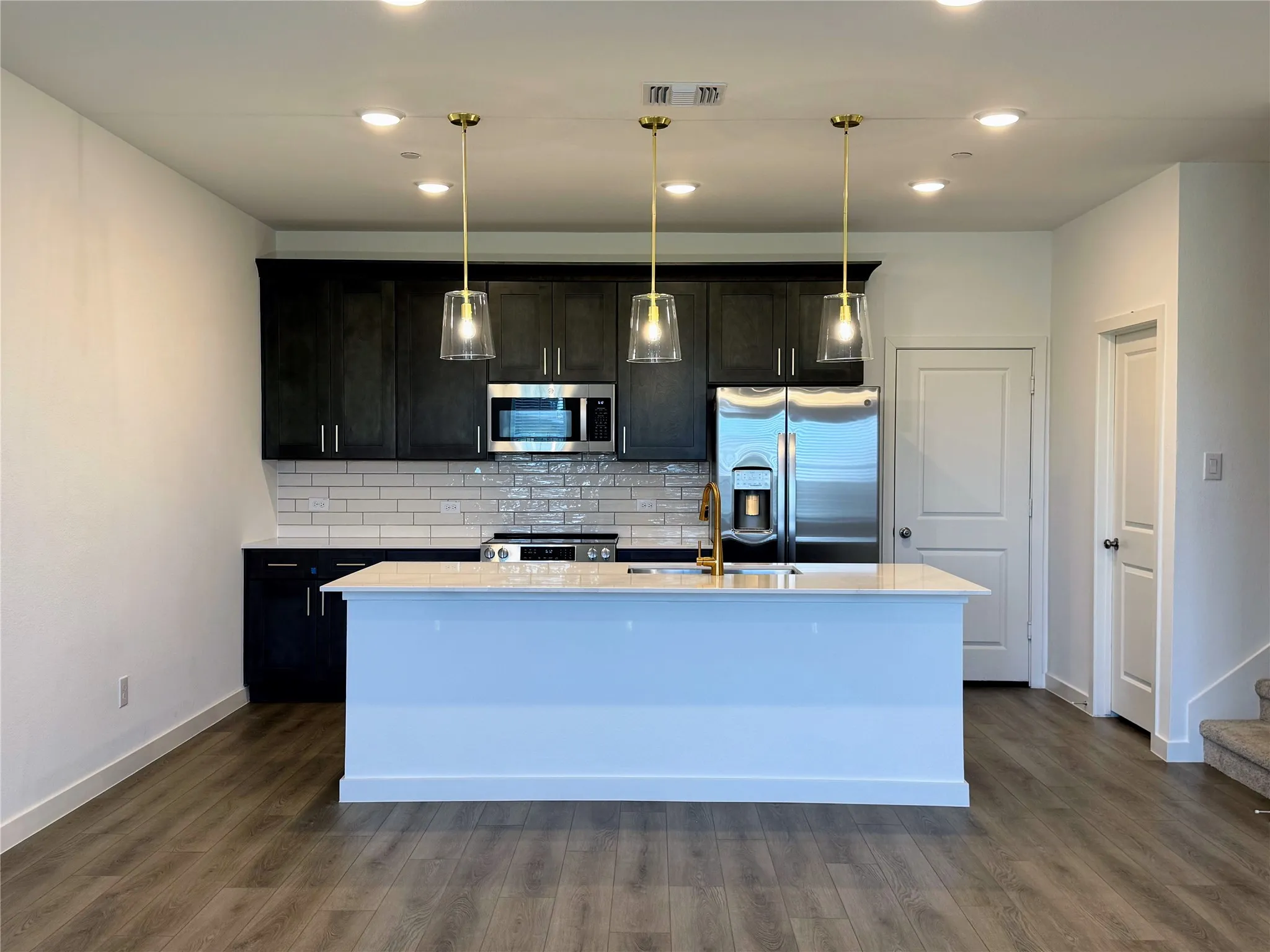 Open-concept kitchen featuring an island with a light-toned countertop, dark cabinetry, stainless steel appliances, a white subway tile backsplash, and wood-finish flooring