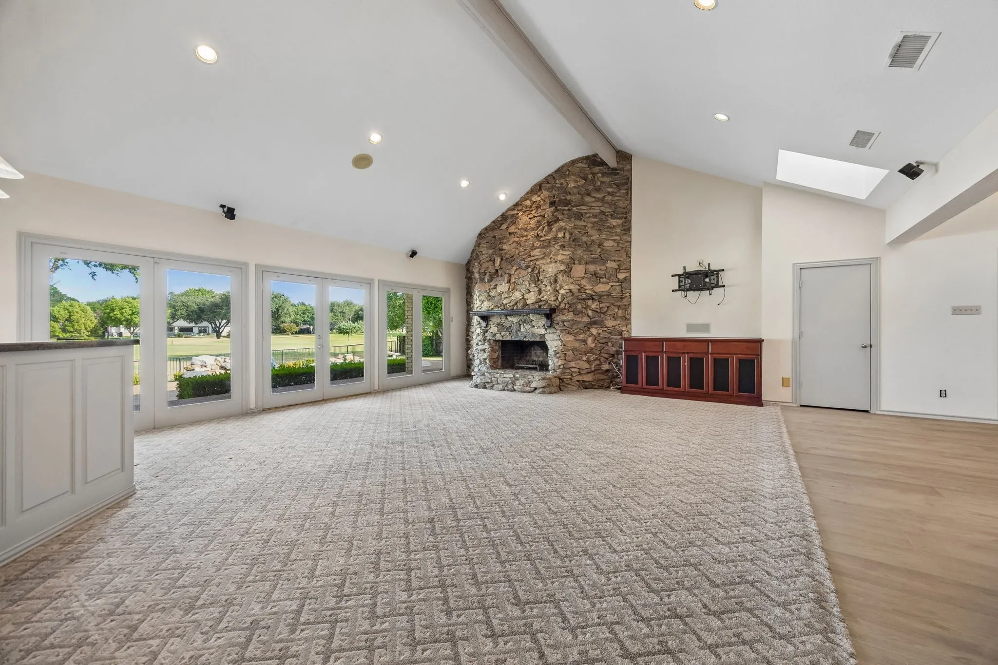 Vaulted ceiling with exposed beam, stone fireplace, multiple glass-paneled doors, integrated skylight, and wood-finish flooring