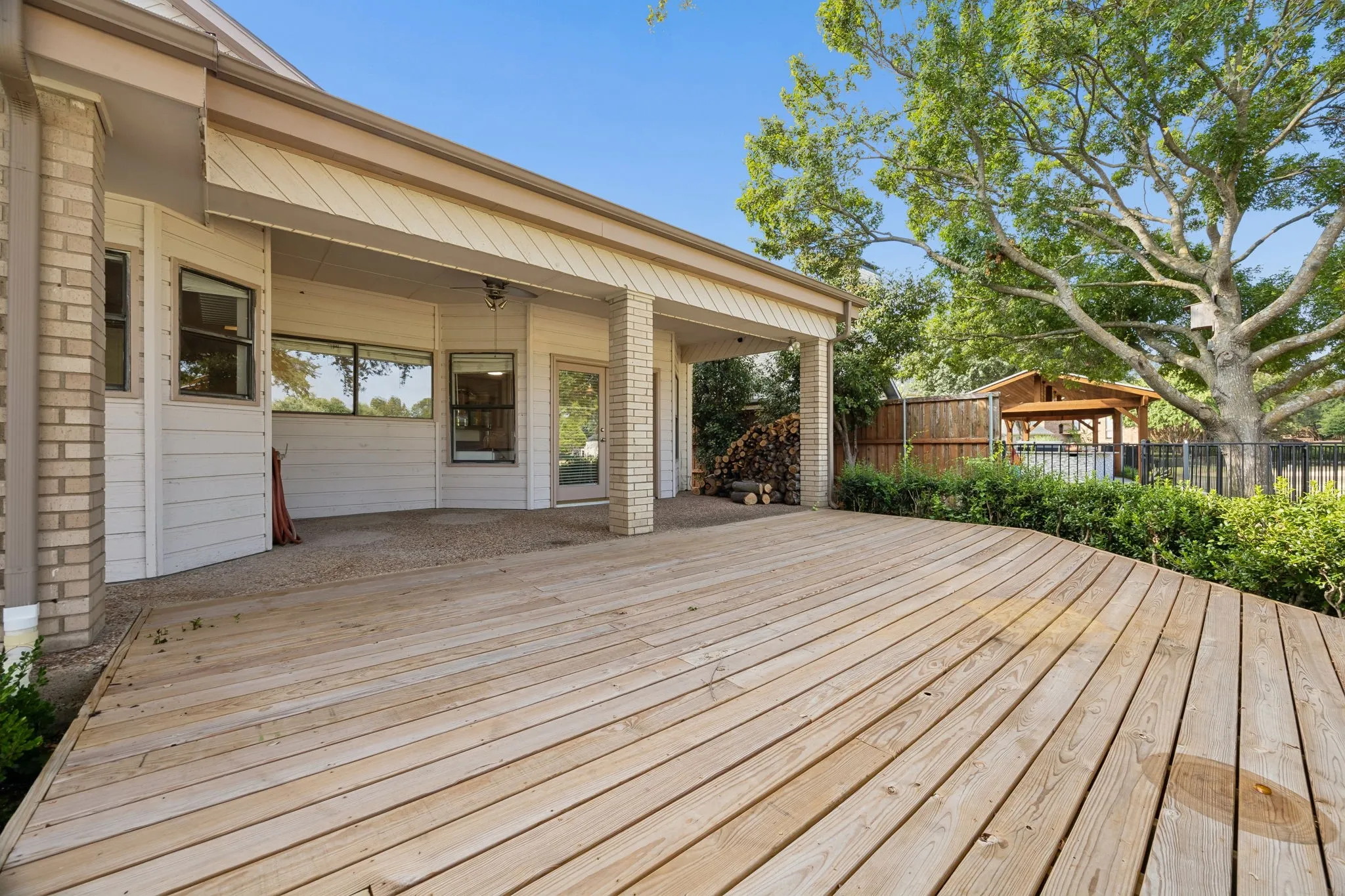 Expansive wood deck with a curved edge, covered patio with brick columns, large windows, and a fenced yard with mature trees