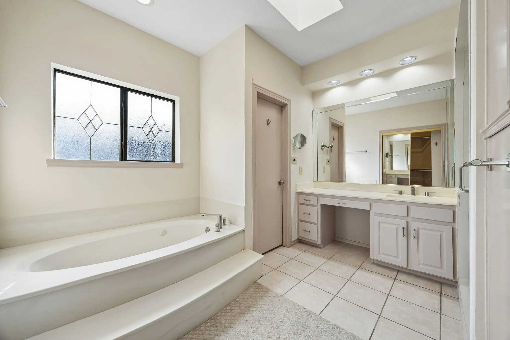 Soaking tub with tiled surround, dual vanity with integrated sink, makeup vanity, large wall mirror, and tiled flooring