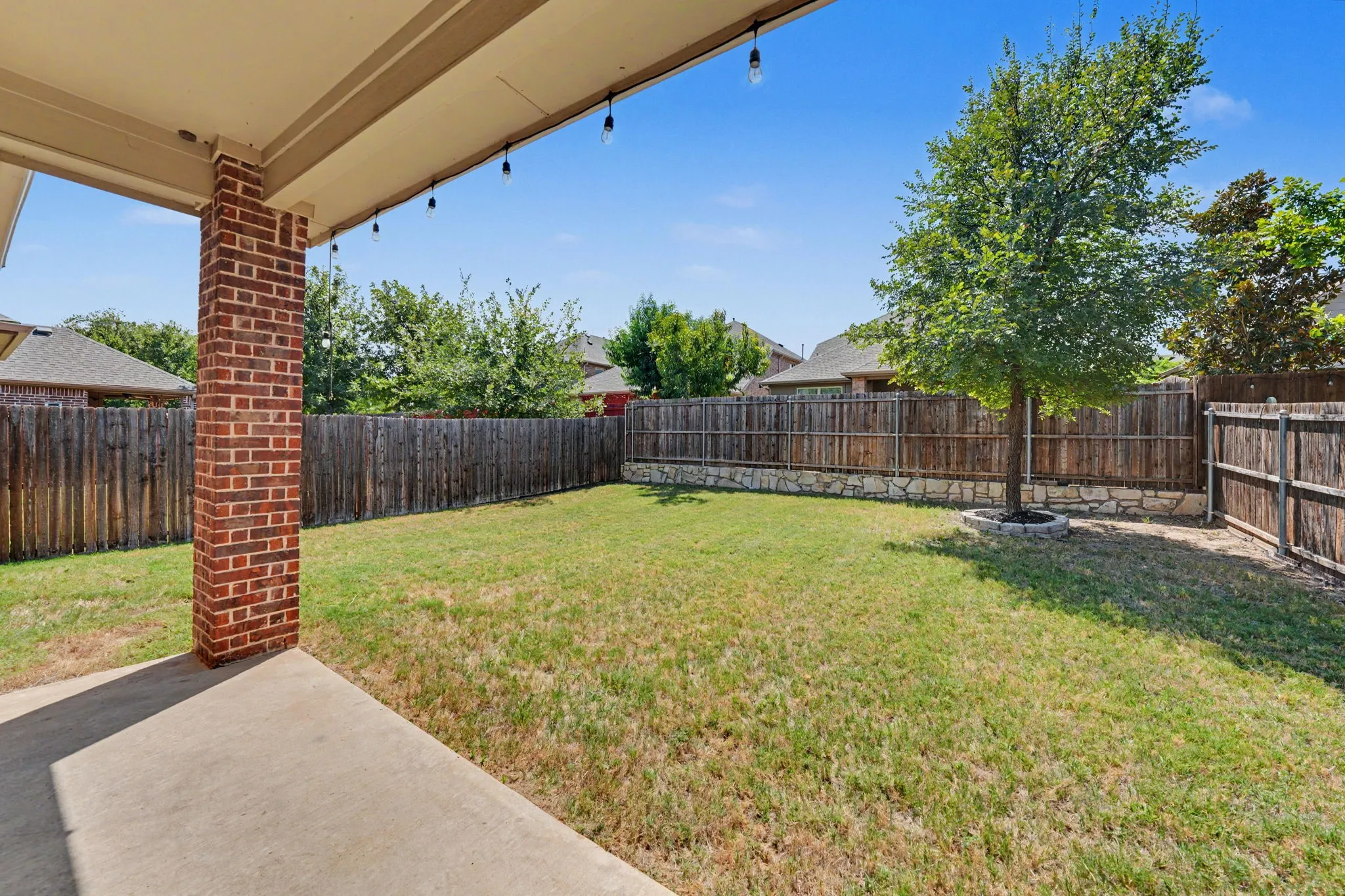 Concrete patio with string lighting, brick column, fenced yard, and stone retaining wall