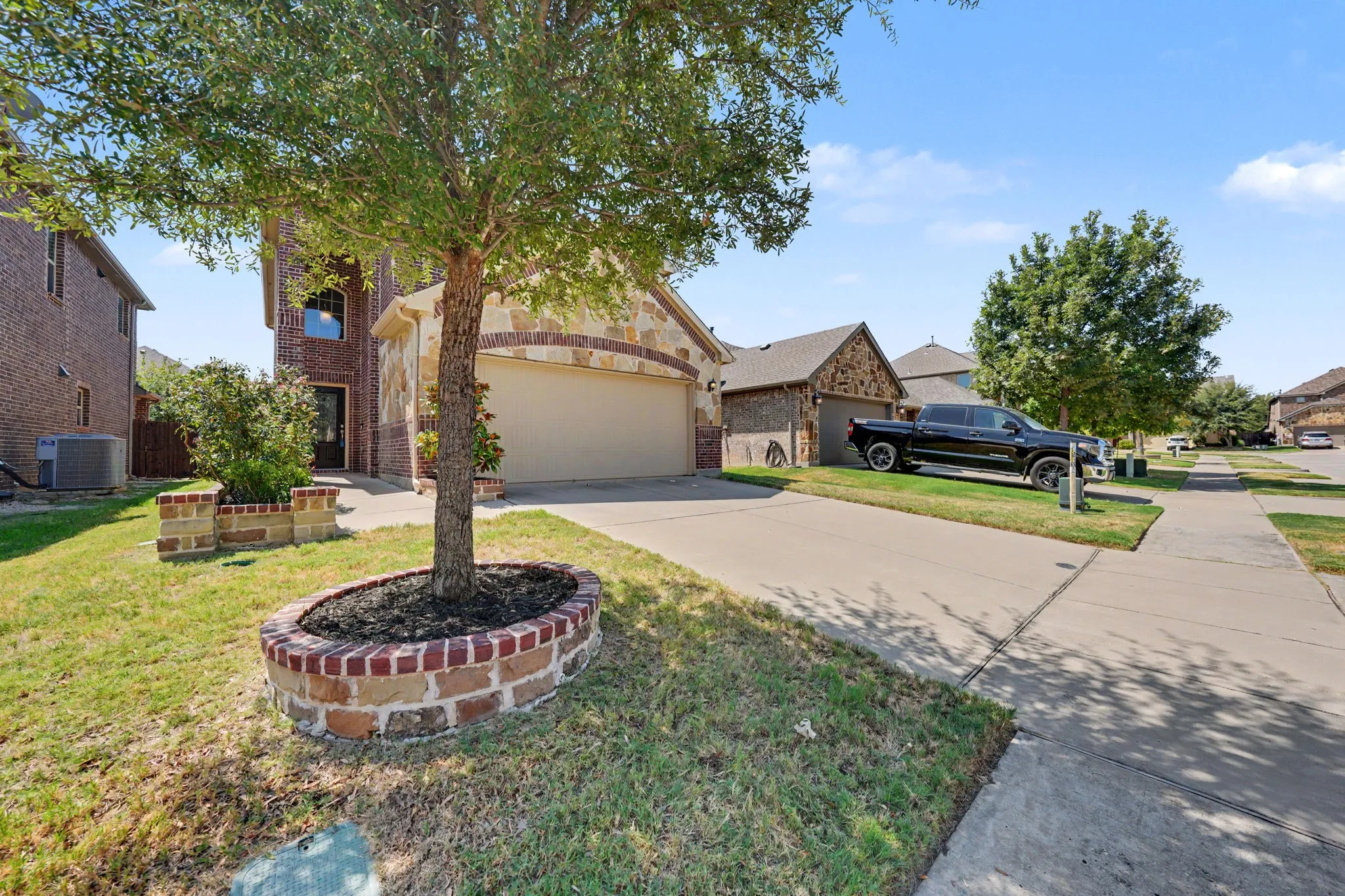 Brick and stone facade, attached garage, well-maintained lawn, and mature tree with a brick planter