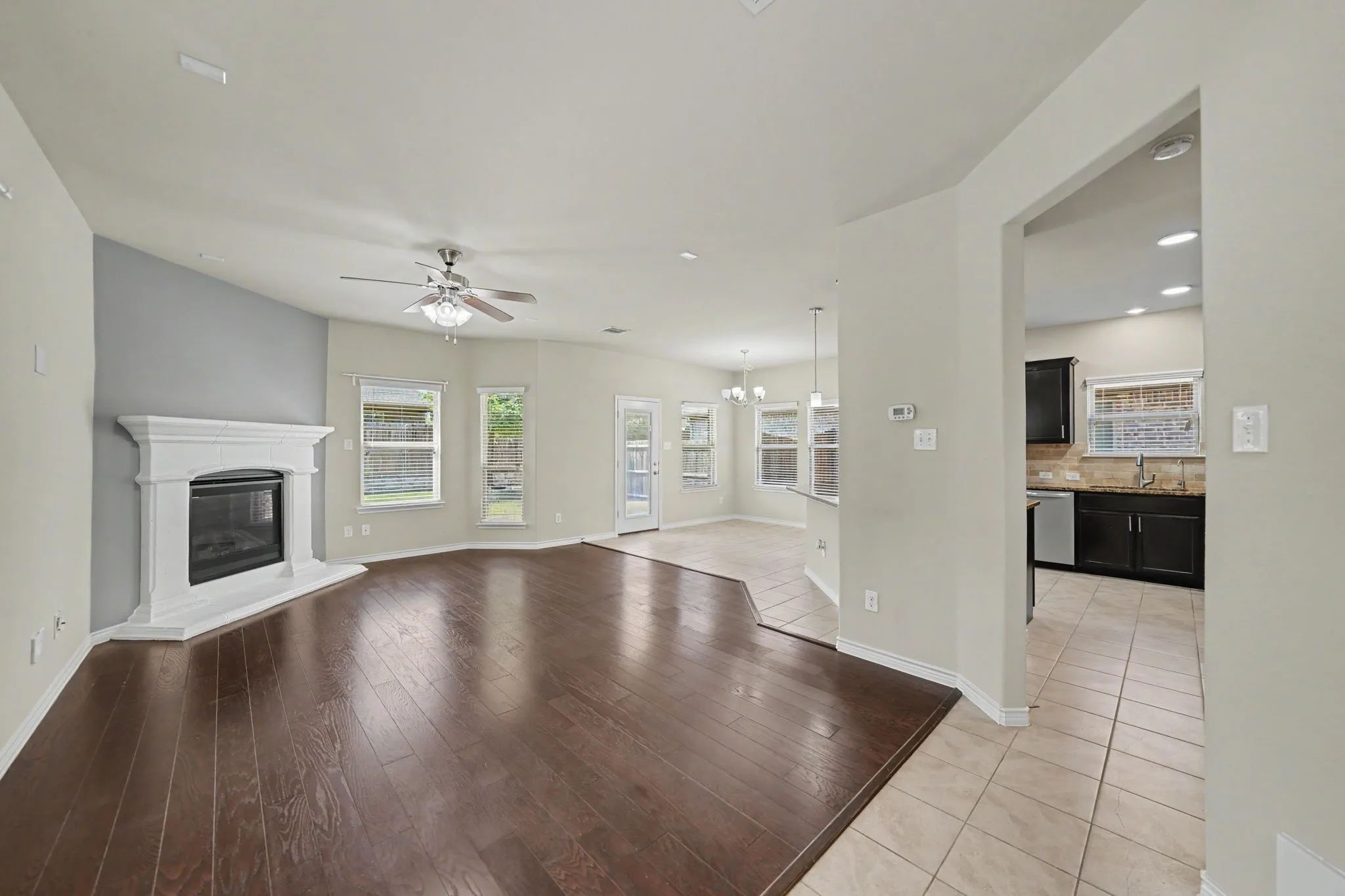 Hardwood-finish flooring, a fireplace with a white mantel, a ceiling fan, multiple windows, and a tiled kitchen with dark cabinetry