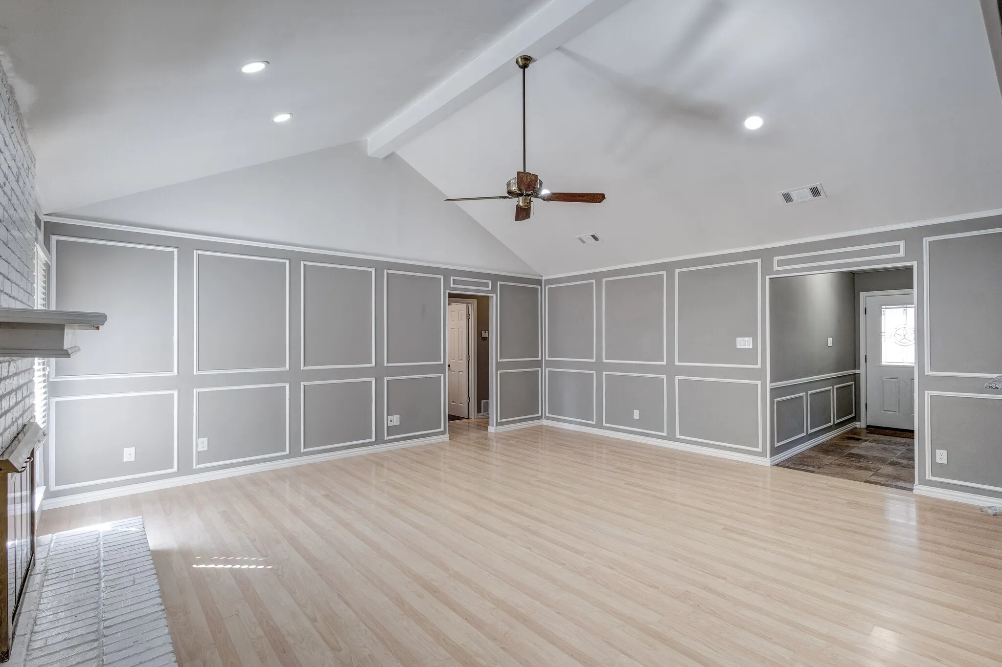 Vaulted ceiling with exposed beam, wood-finish flooring, accent wall paneling, whitewashed brick fireplace, and ceiling fan