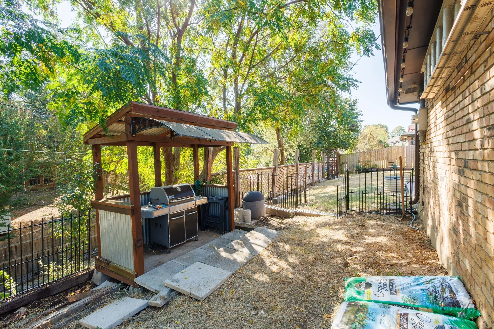 Brick exterior with downspout, covered outdoor cooking area, wood-frame gazebo with corrugated roof, and wrought iron fencing