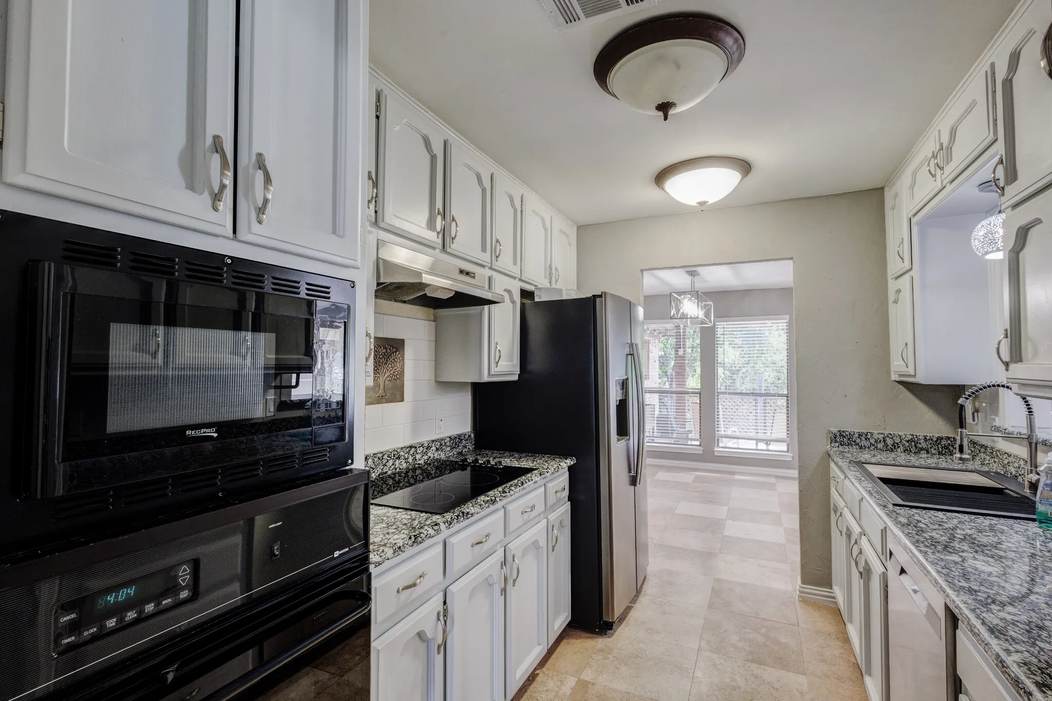 White cabinetry with silver-tone hardware, dark countertop with integrated sink, built-in microwave and oven, stainless steel refrigerator, and tiled flooring