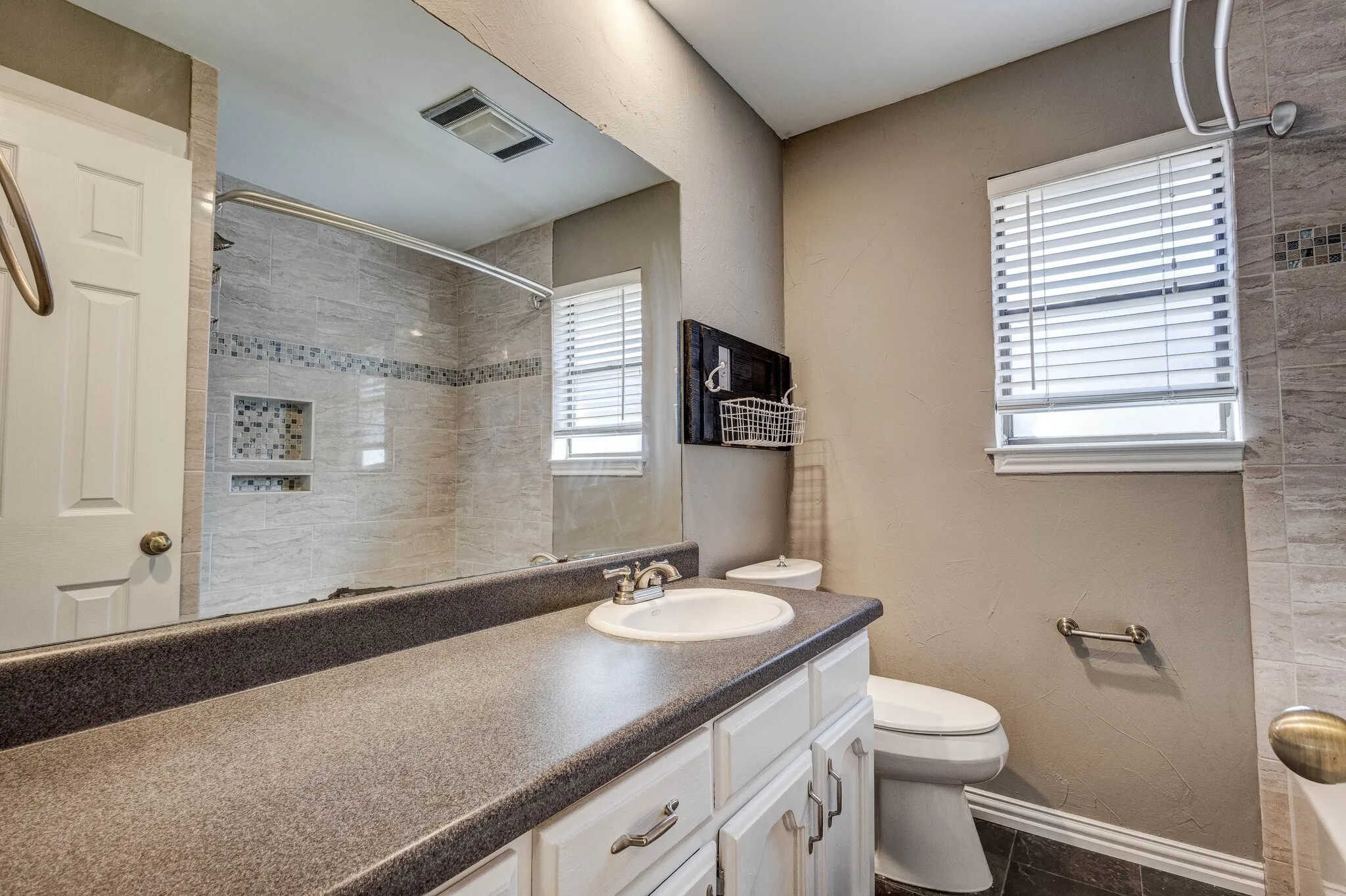 Bathroom featuring a vanity with a white basin and dark countertop, white cabinetry with silver hardware, a toilet, a window with blinds, and a shower with tile surround and built-in niches