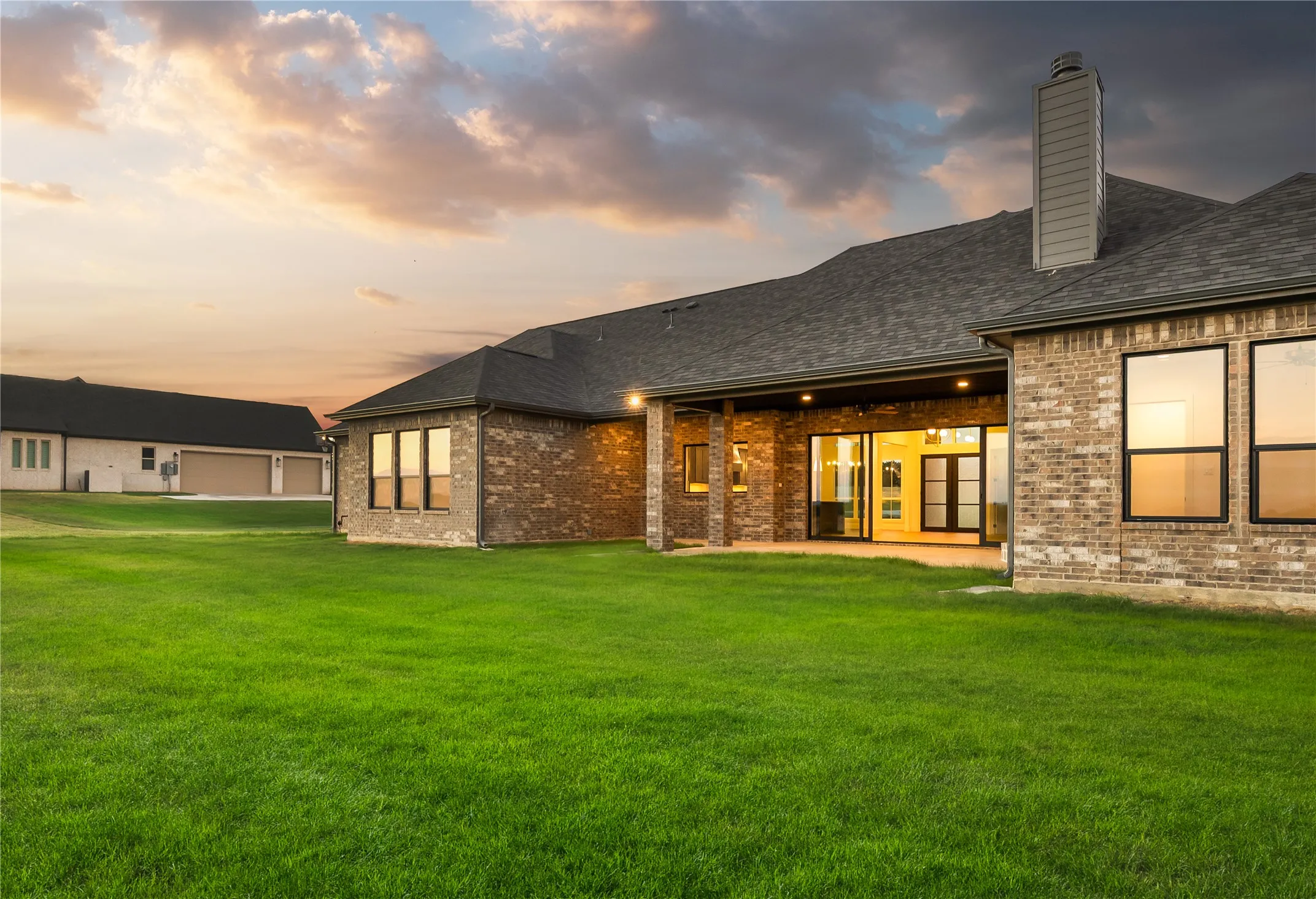 Expansive green lawn with a brick exterior, multiple large windows, a covered patio, and sliding glass doors