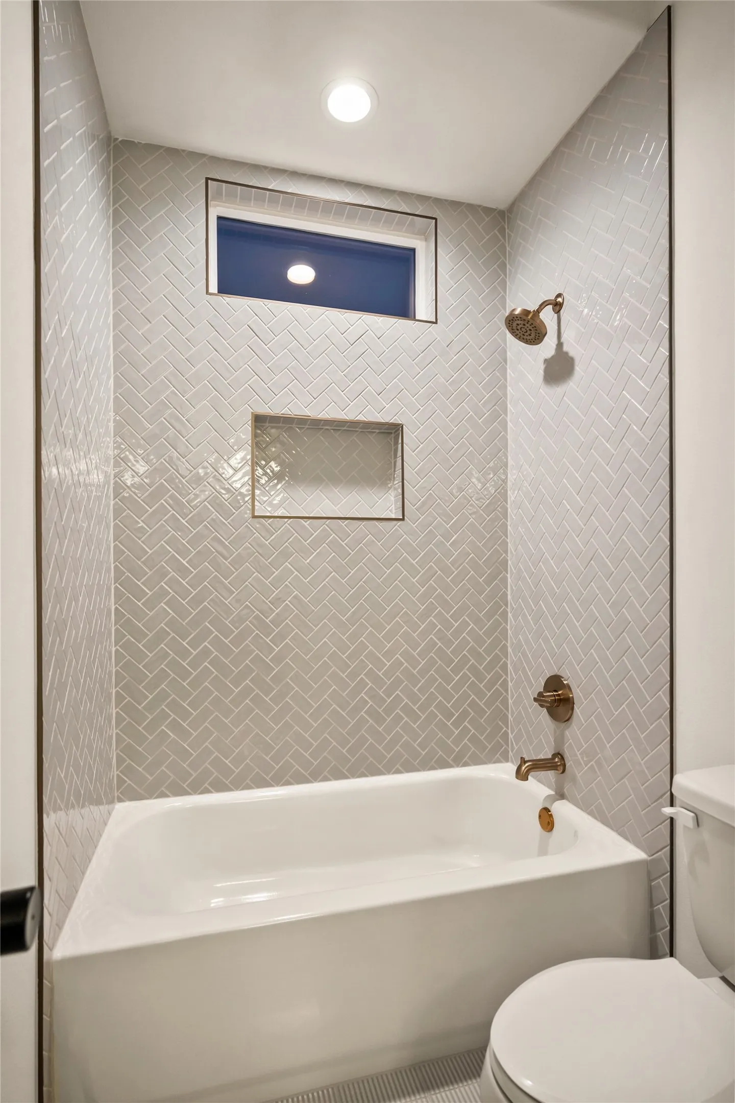 Bathtub with herringbone tile surround, bronze-finish fixtures, and a recessed window