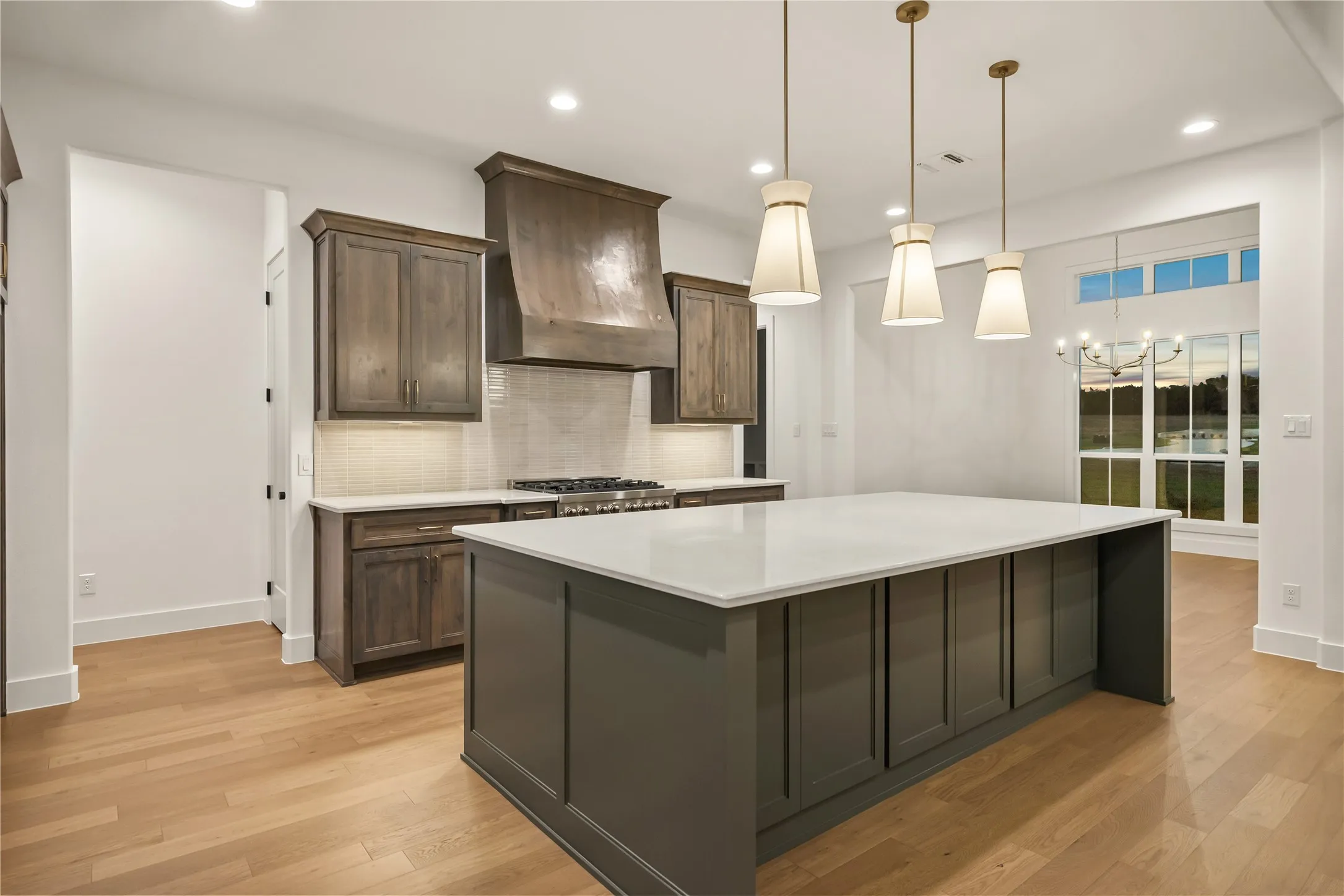 Spacious kitchen featuring an expansive central island with a light-toned countertop, wood-finish flooring, recessed lighting, and a window framing outdoor scenery