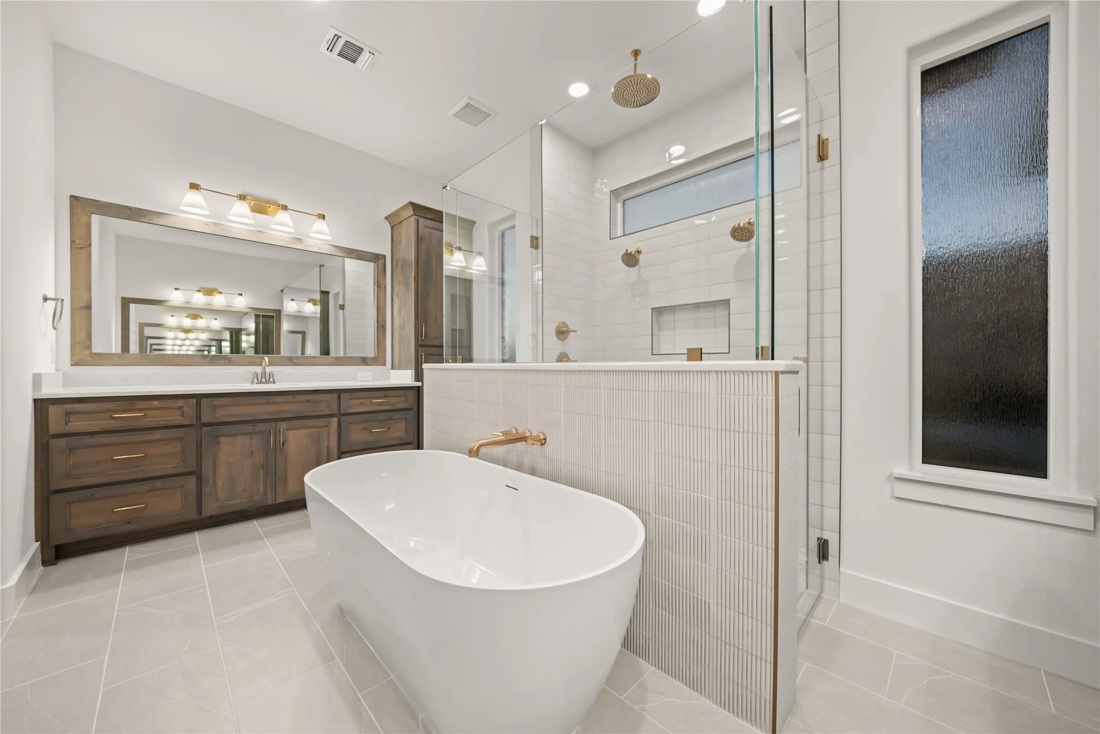 Freestanding soaking tub with gold-finish fixtures, a spacious walk-in shower with dual shower heads, a long vanity with wood-finish cabinetry, a large framed mirror, and light tile flooring