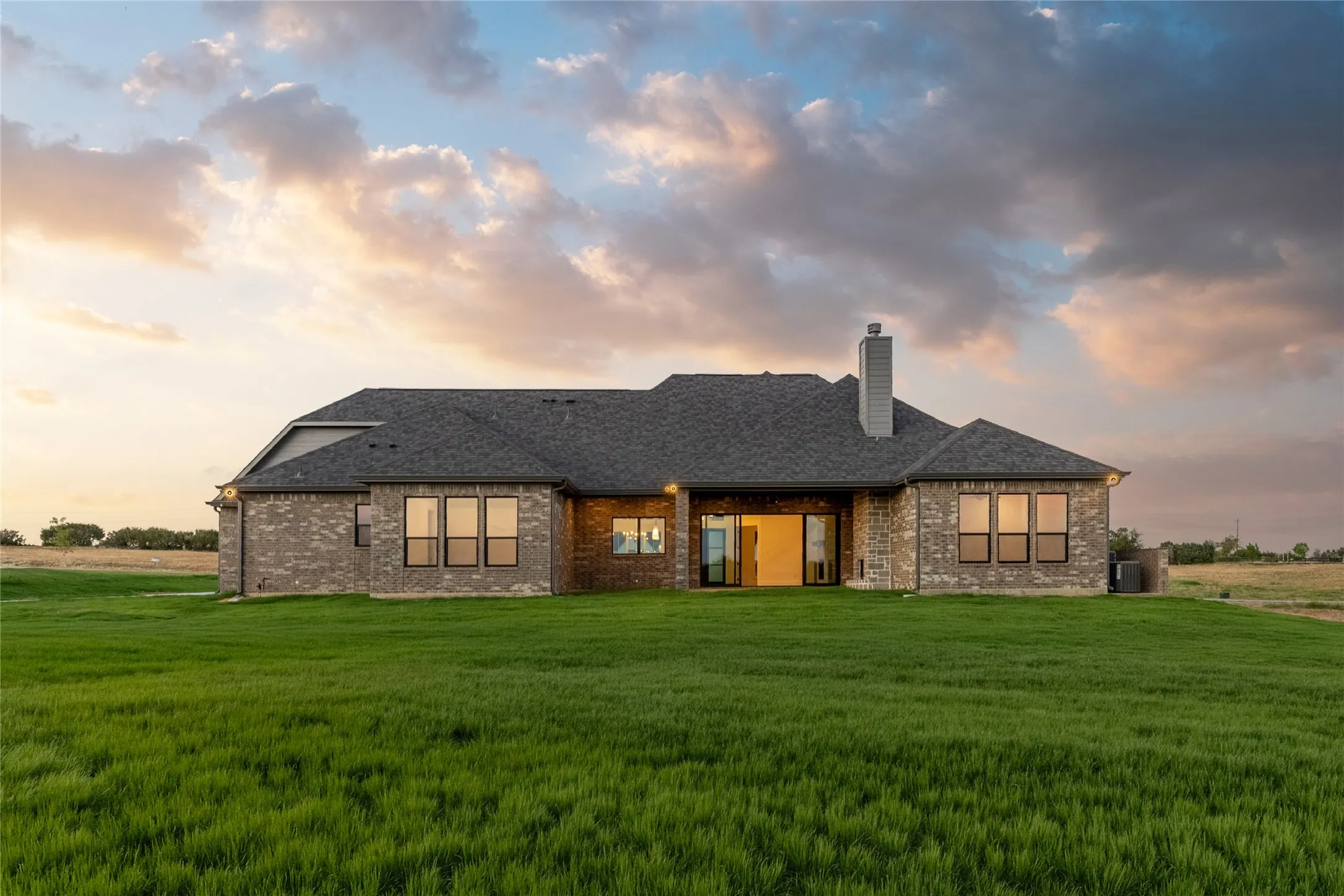 Expansive green lawn surrounds the rear elevation, featuring a brick exterior, multiple windows with dark frames, a prominent chimney, and a sliding glass door