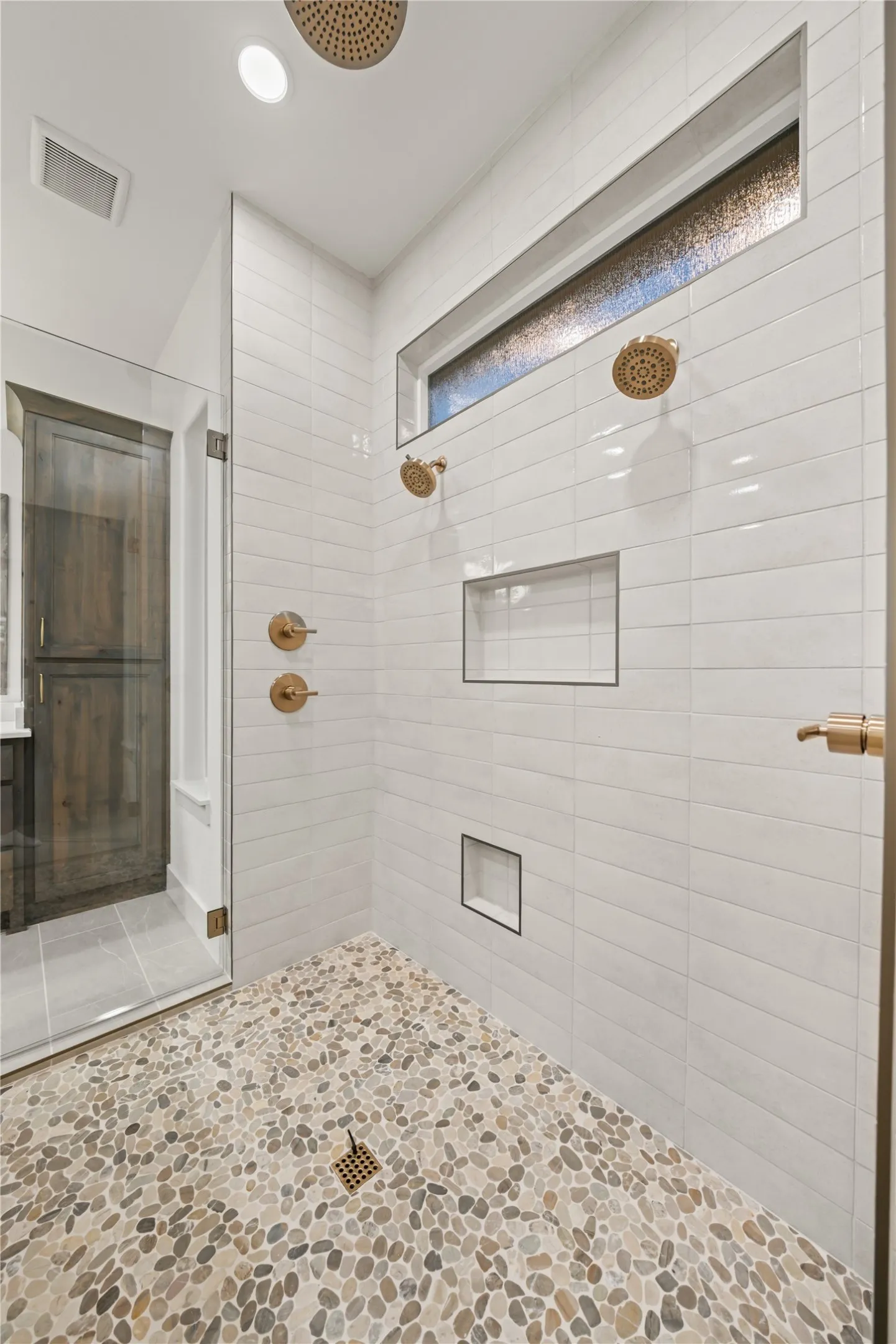 Spacious walk-in shower with pebble tile flooring, white subway tile walls, dual shower heads, and built-in niches