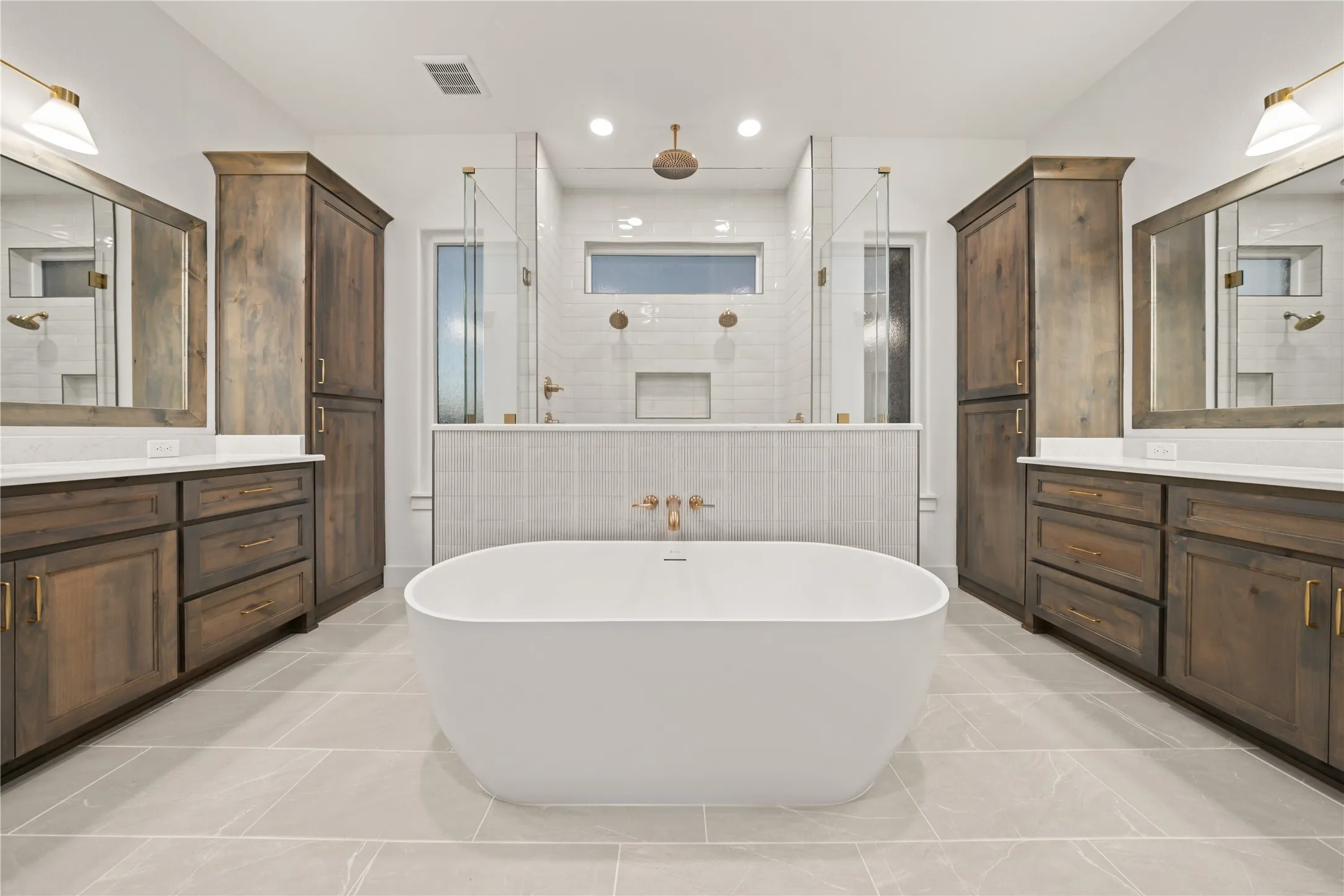 Freestanding soaking tub, dual vanities with wood-finish cabinetry, frameless glass shower enclosures, and light-toned tile flooring