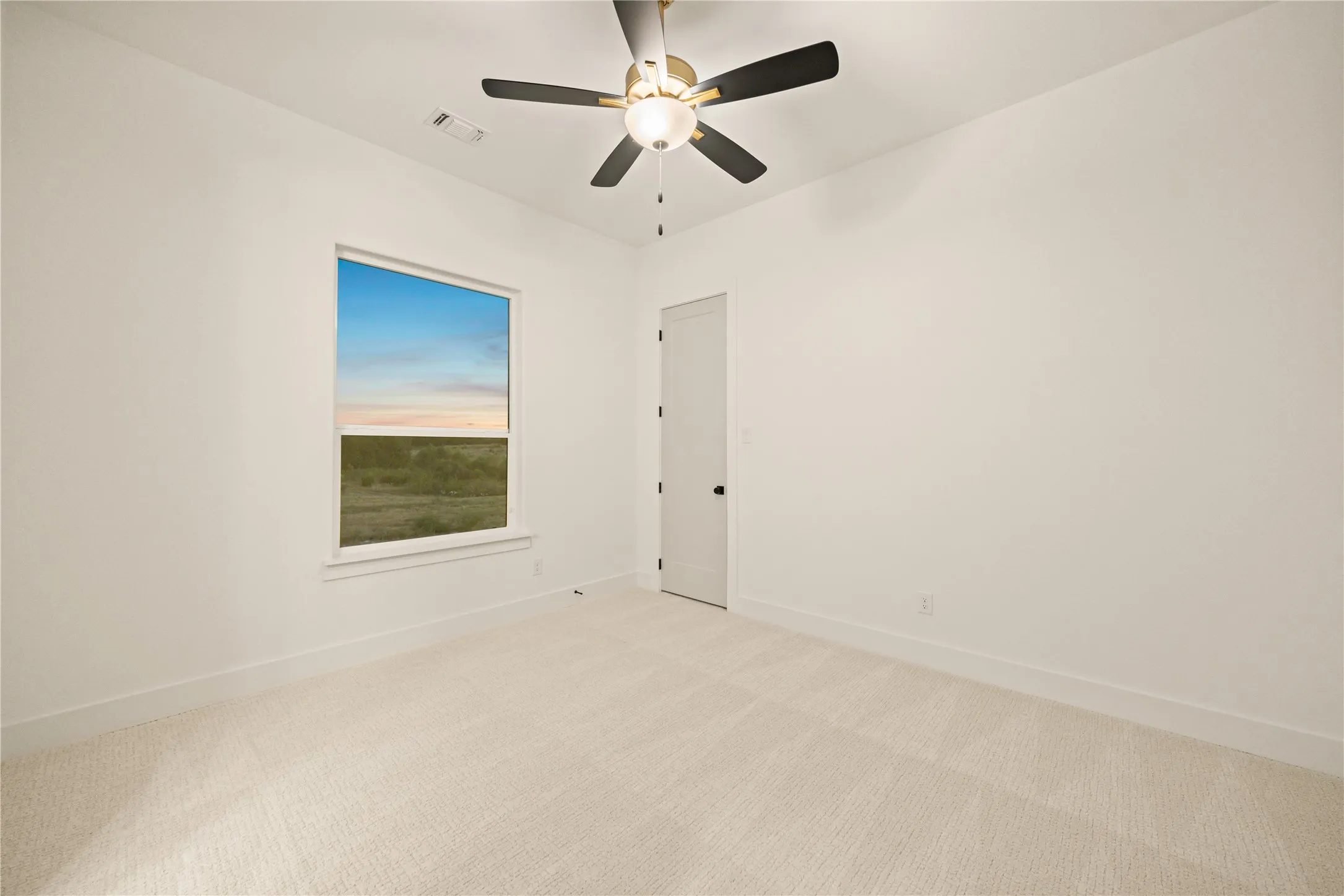 White walls, light-colored carpeting, a window with an outdoor landscape, and a ceiling fan with light fixture