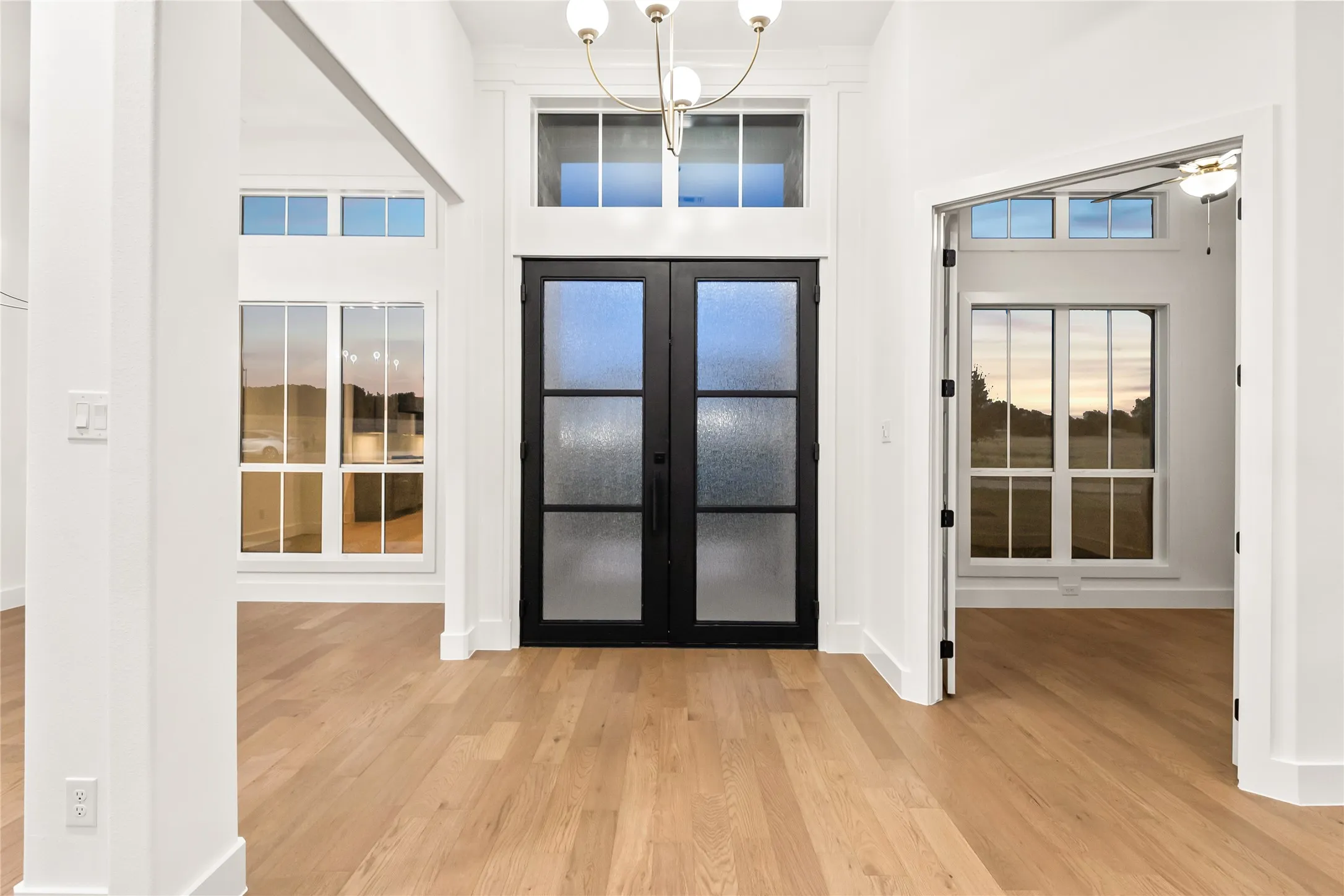 Wood-finish flooring, white walls, black double-entry doors with obscured glass panels, a brass chandelier, and transom windows