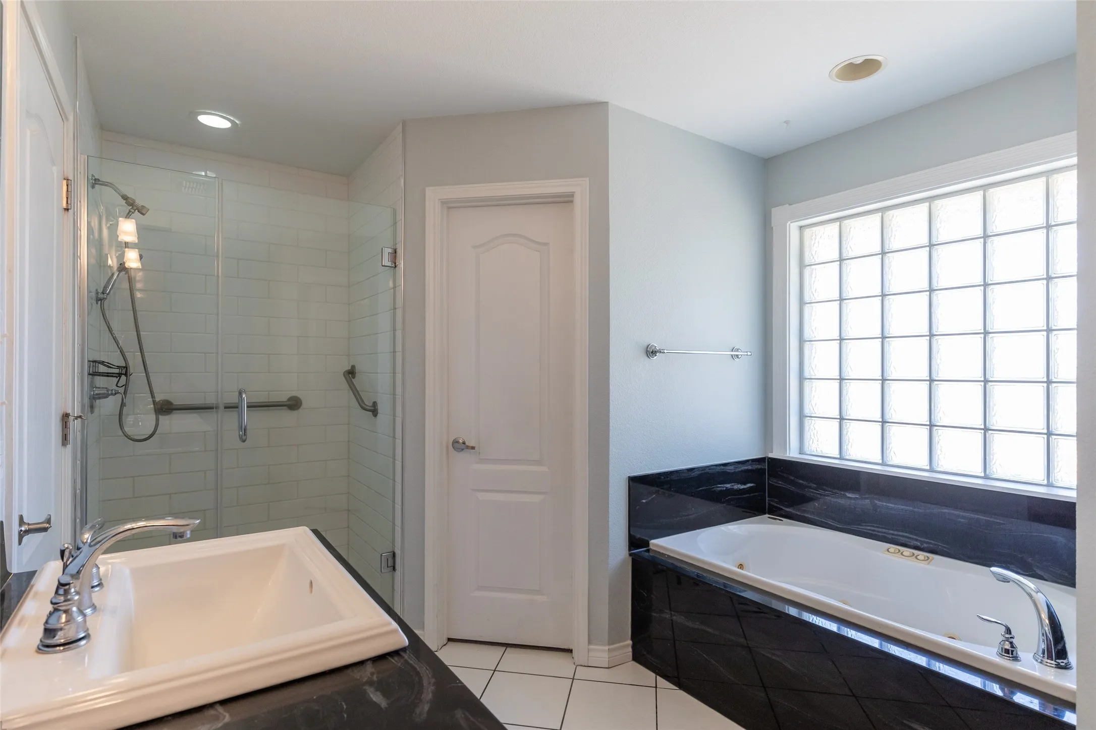 Frameless glass shower enclosure with subway tile surround, integrated soaking tub with dark stone-finish surround, single vanity with dark stone-finish countertop and undermount sink, and glass block window