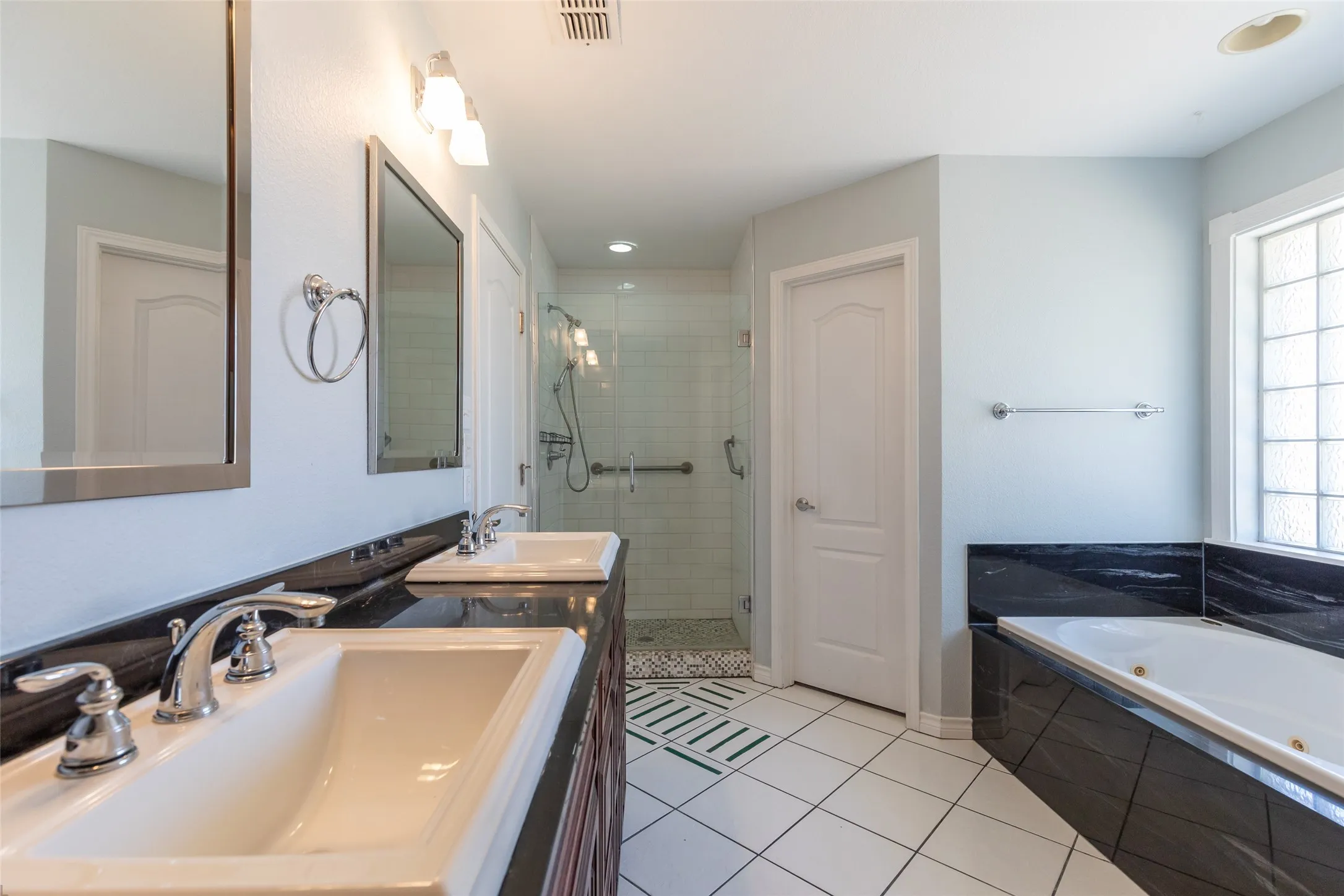 Dual vanity with integrated sinks, dark stone countertops, separate glass-enclosed shower, white tile flooring, and a built-in jetted tub with dark stone surround