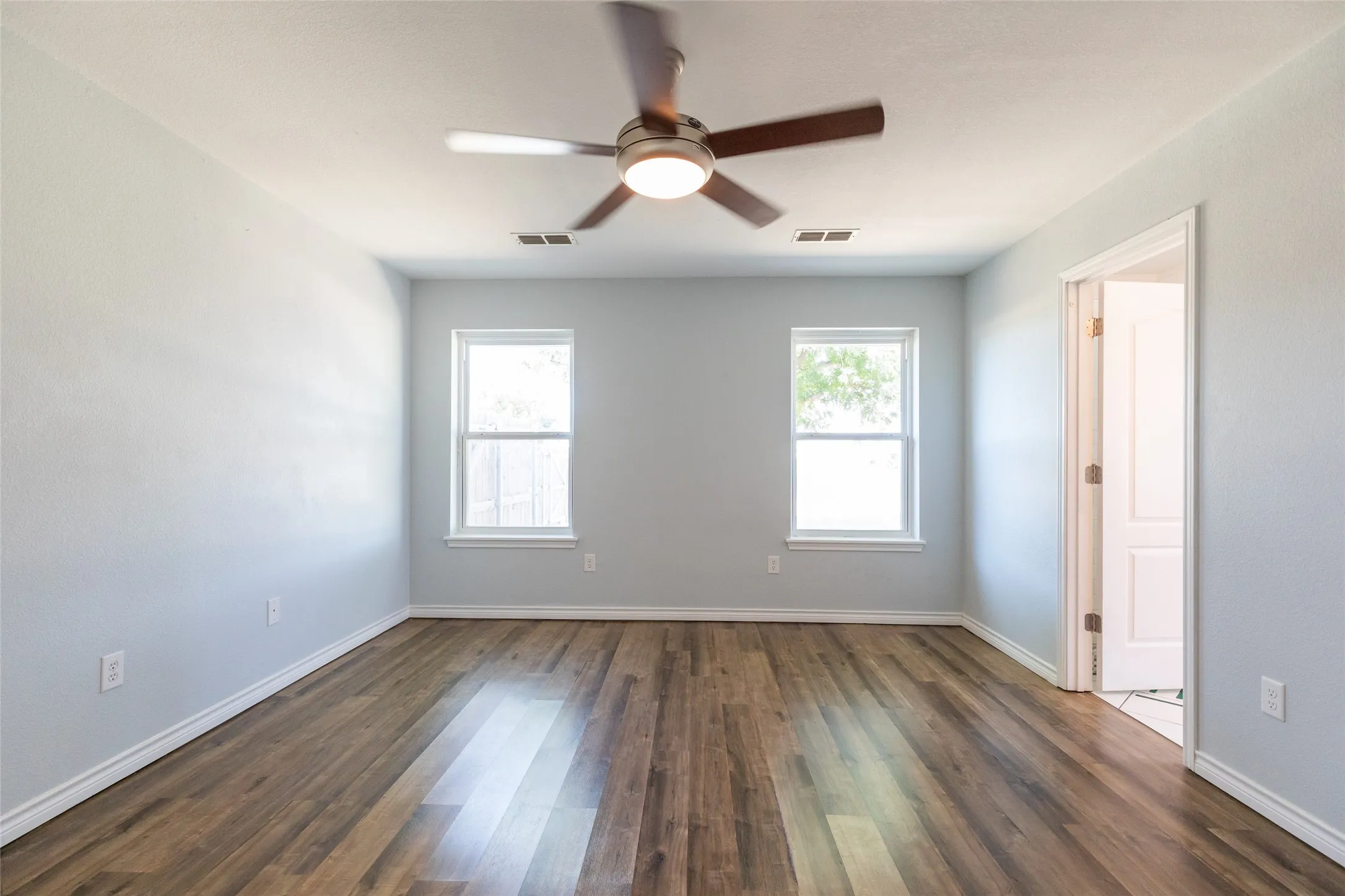 Wood-finish flooring, two large windows, overhead ceiling fan with integrated lighting, white trim, and a paneled interior door