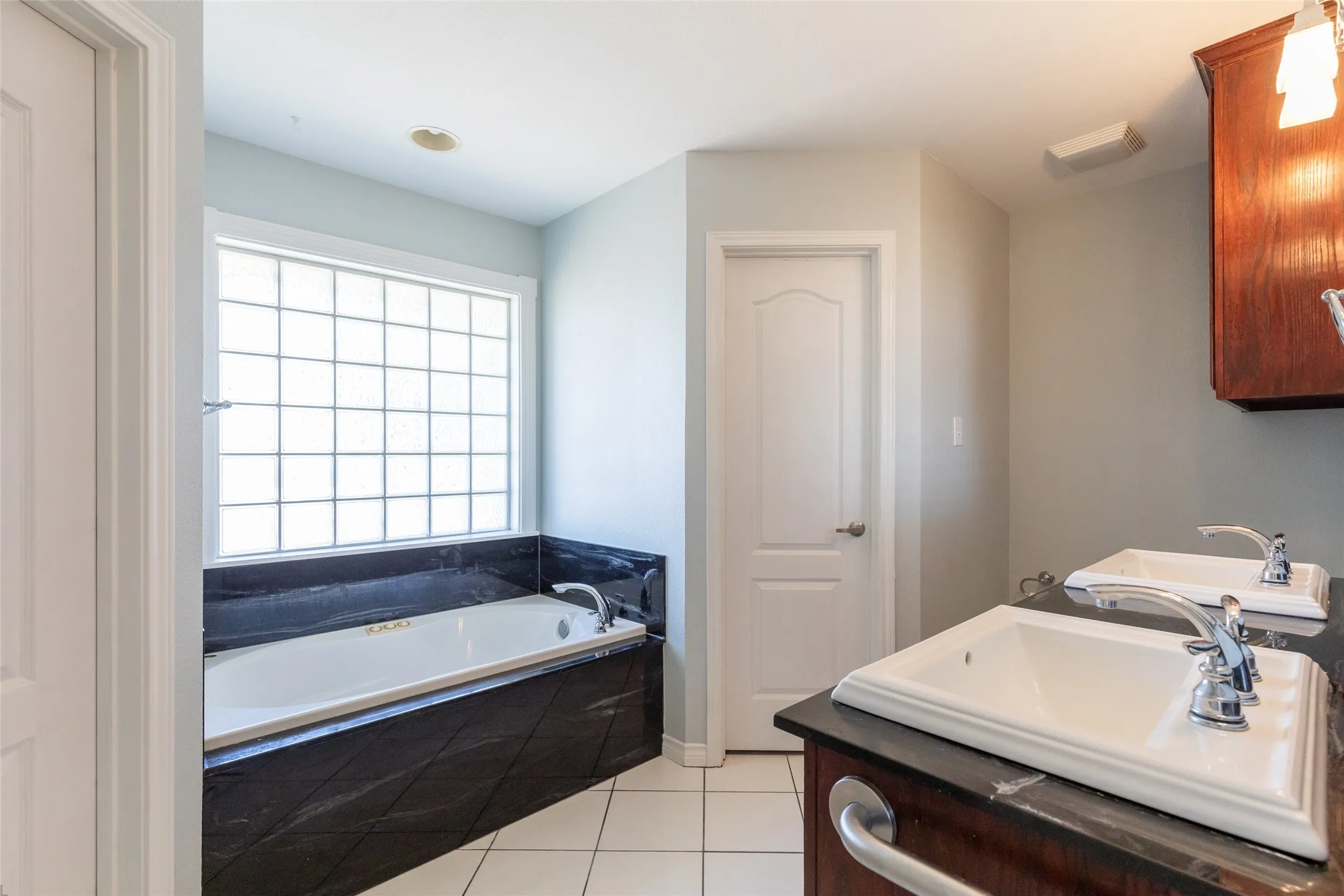 Built-in soaking tub with dark tile surround, glass block window, dual vanity sinks, and wood-finish cabinetry