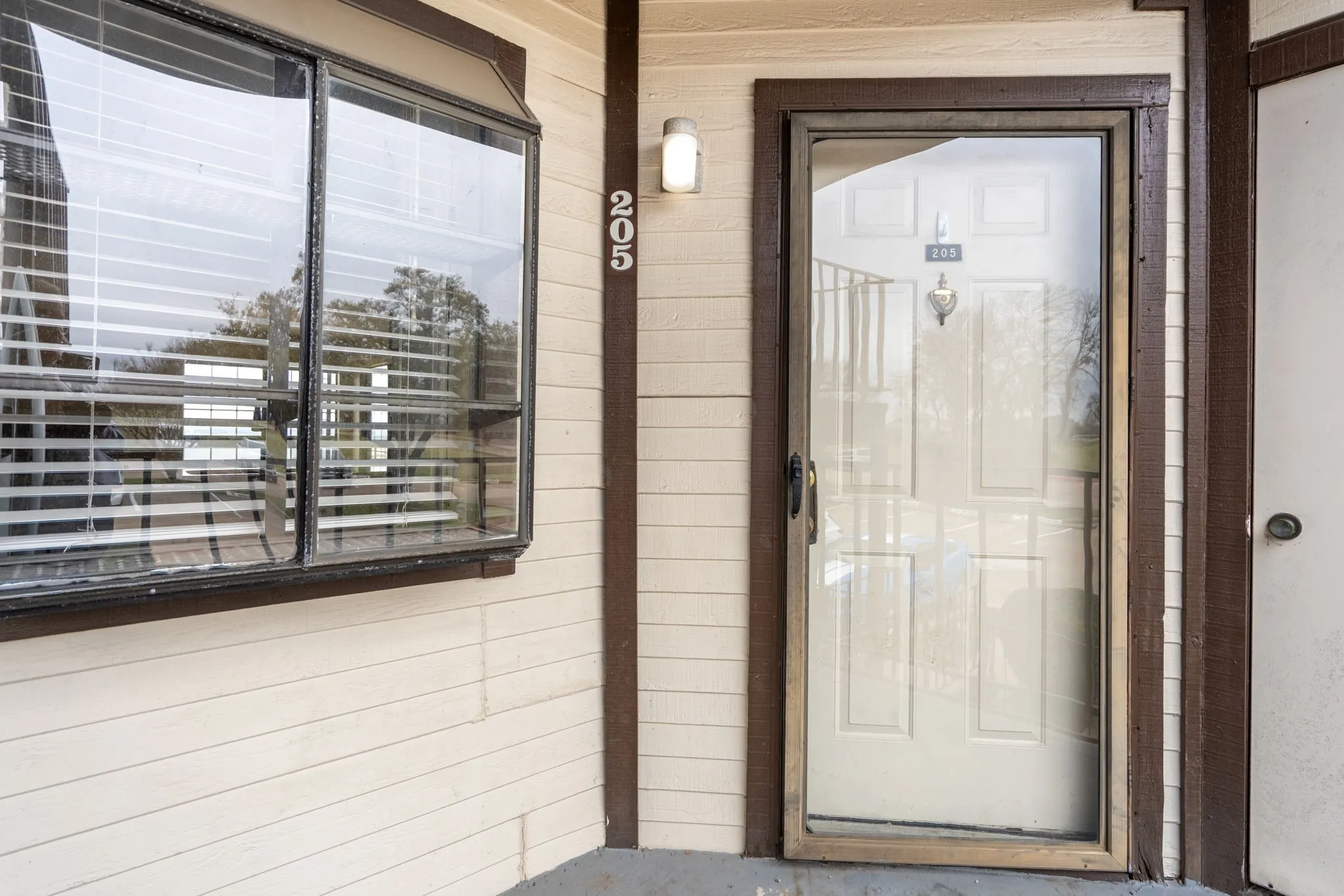Bay window with blinds, light-colored horizontal siding, dark trim, wall-mounted light fixture, and unit number 205