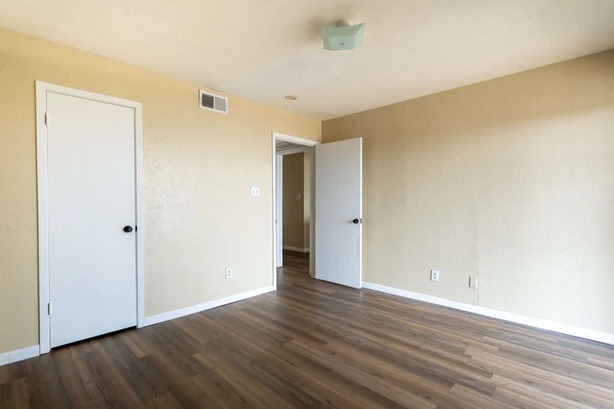 Wood-finish flooring throughout, multiple white doors with black hardware, neutral wall paint, and overhead lighting fixture