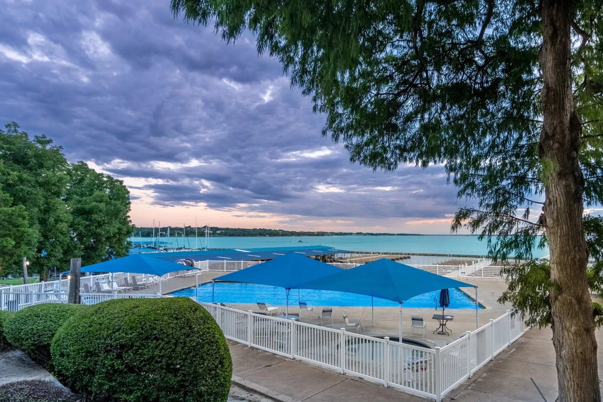 Fenced swimming pool area with blue shade structures, expansive water backdrop, and surrounding green landscaping