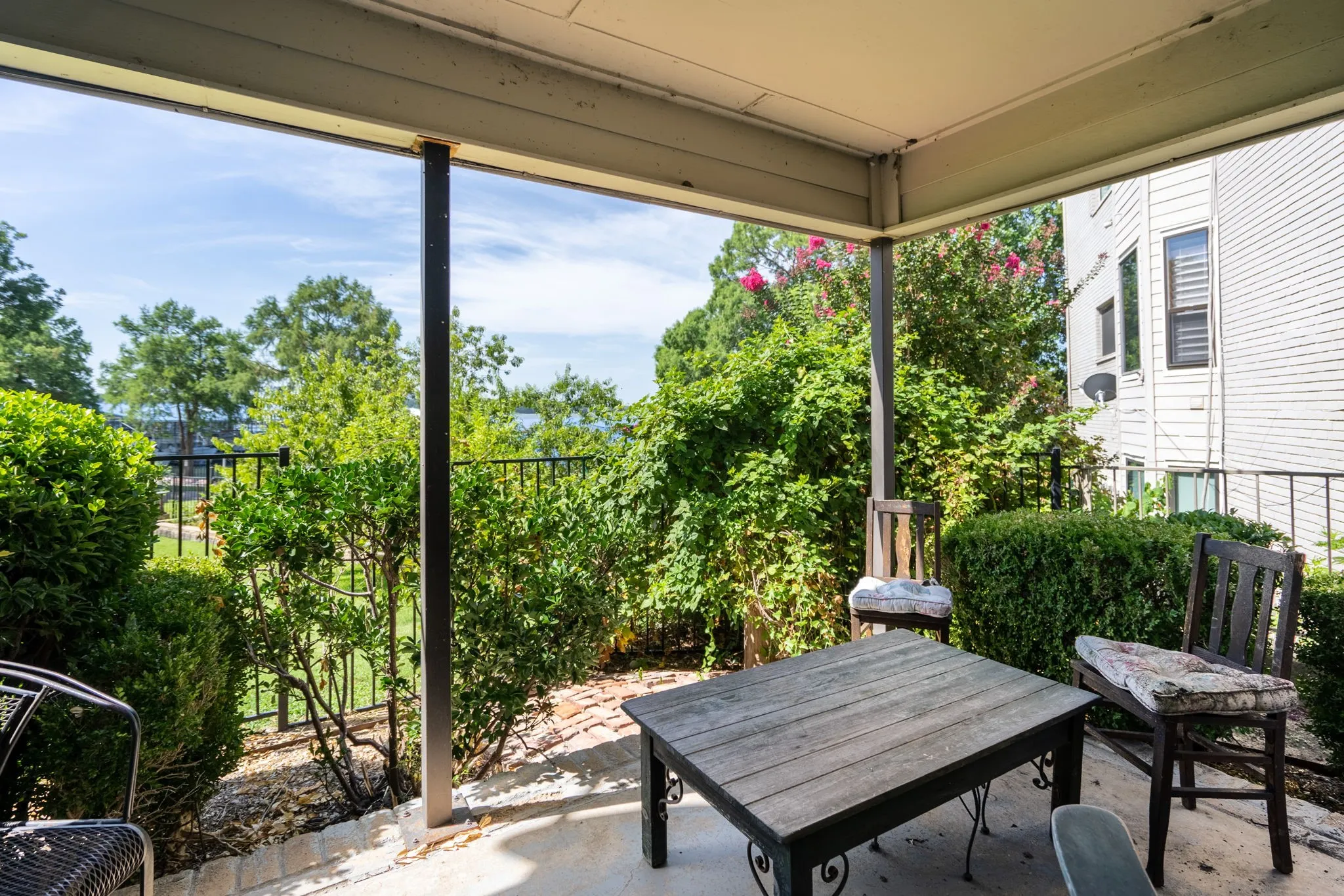 Covered patio with concrete flooring, framed by mature shrubs and a black metal fence