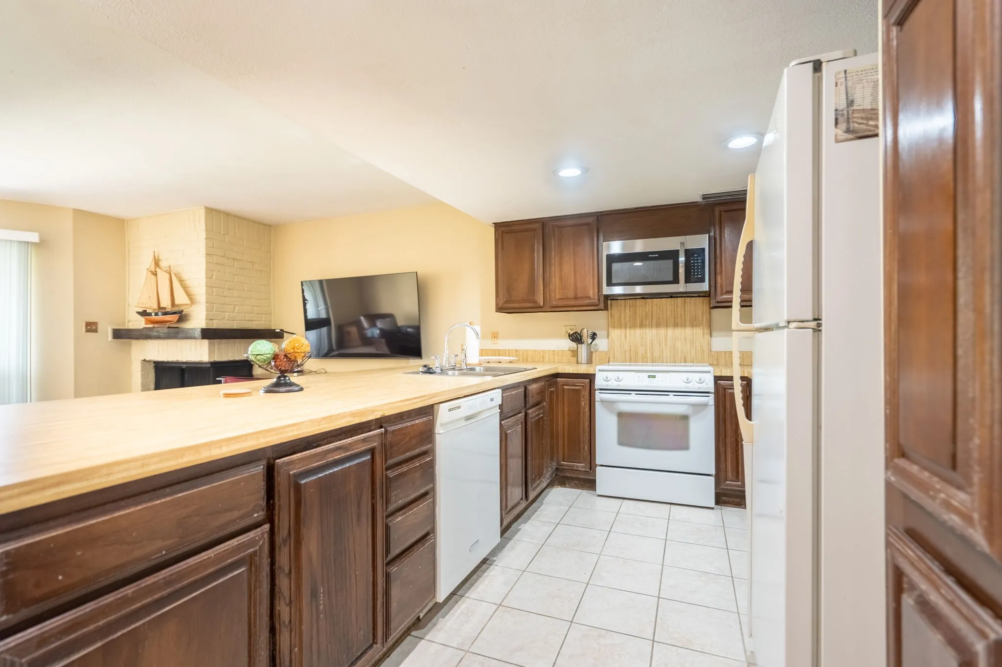 Open-concept kitchen featuring wood-finish cabinetry, light-toned countertops, white tile flooring, and integrated white appliances