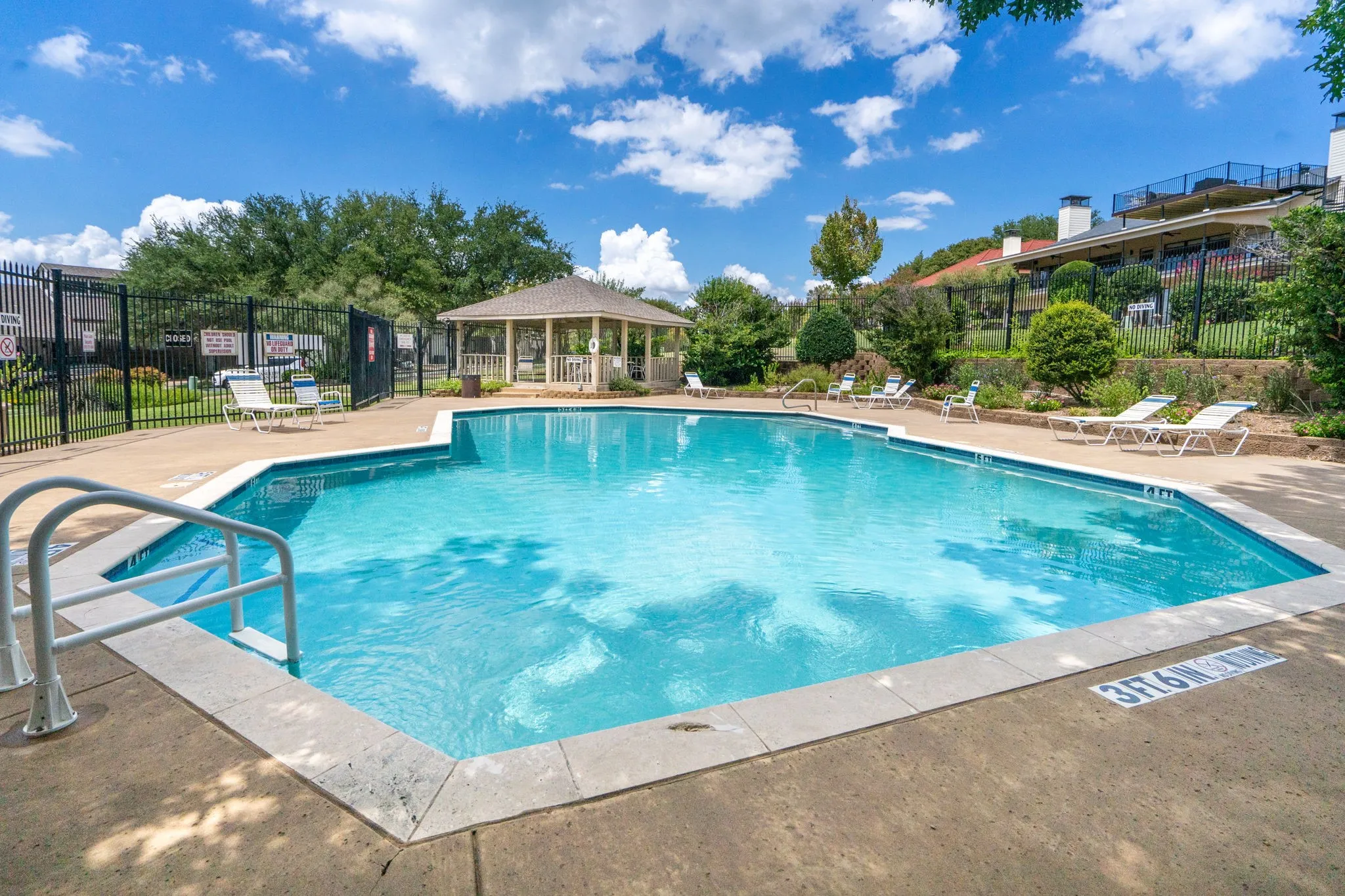 In-ground swimming pool with concrete decking, surrounded by a black metal fence and a nearby gazebo