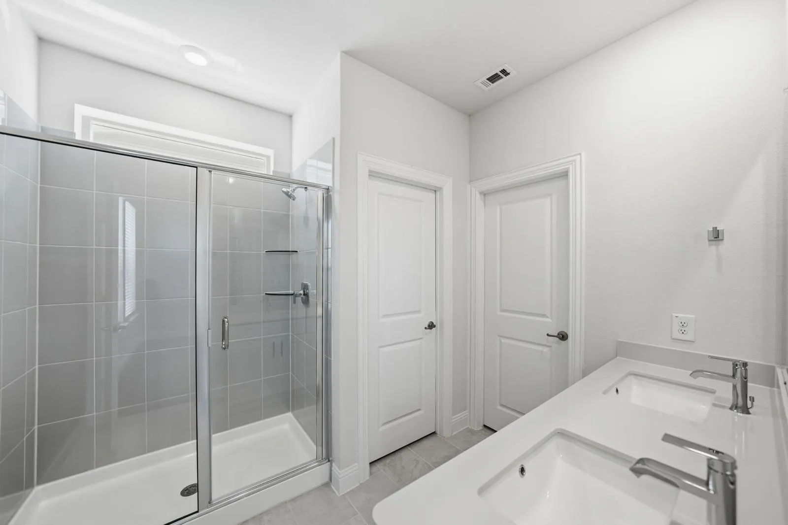 Dual sinks with chrome fixtures, a spacious walk-in shower with glass enclosure, light gray tiled shower walls, and white countertops