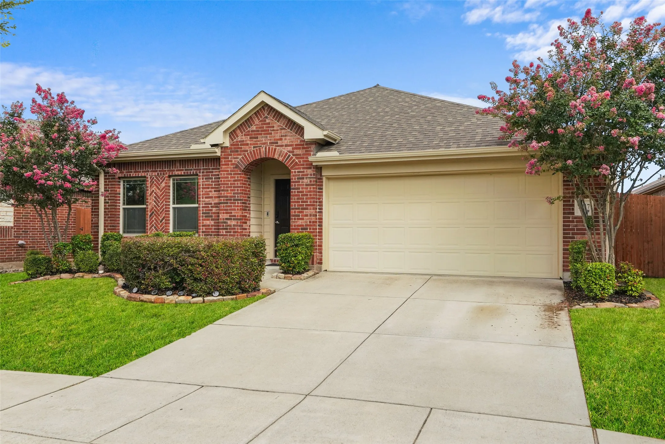 Brick facade with an arched entry, attached garage, concrete driveway, and manicured landscaping featuring flowering trees