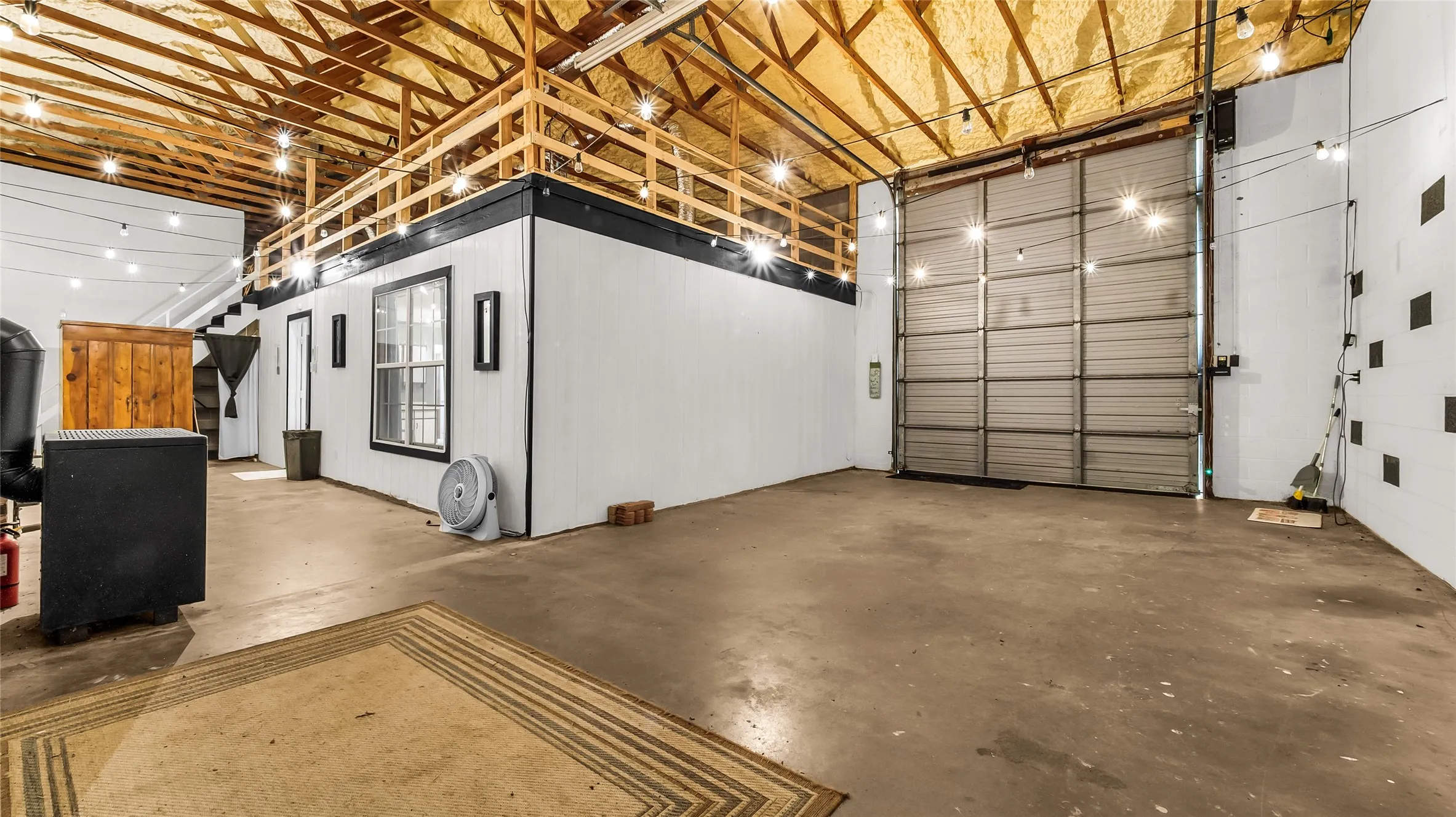 Exposed wood beam ceiling, concrete flooring, upper-level mezzanine with wood railing, interior wall with window, and large roll-up door
