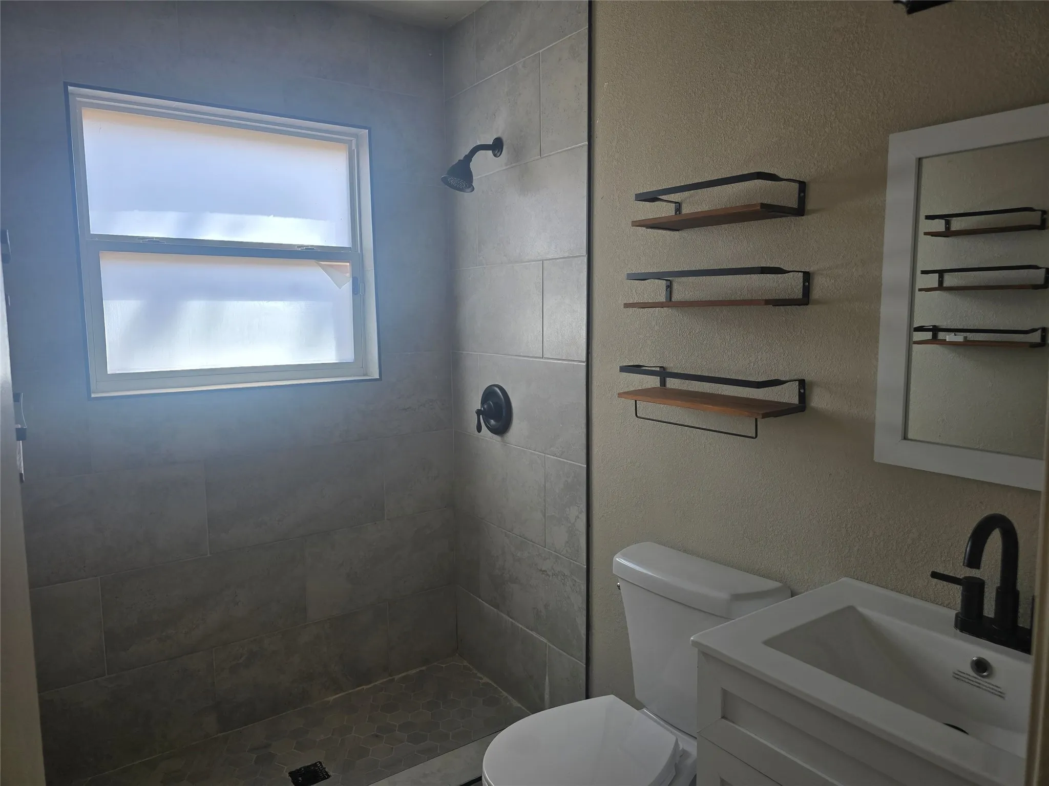 Tiled shower with dark bronze fixtures, white vanity with integrated sink, white framed mirror, and floating wood-finish shelves
