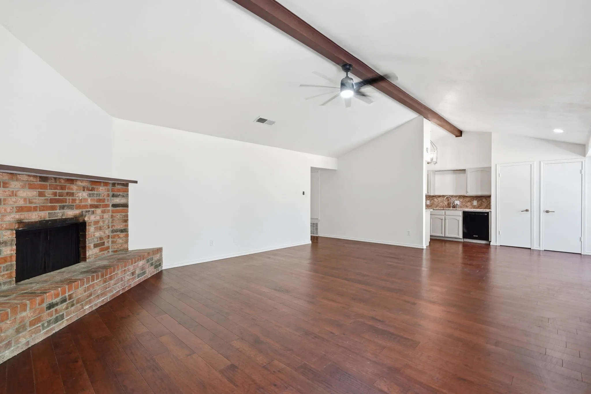 Brick fireplace with hearth, vaulted ceiling, exposed beam, ceiling fan, and wood-finish flooring