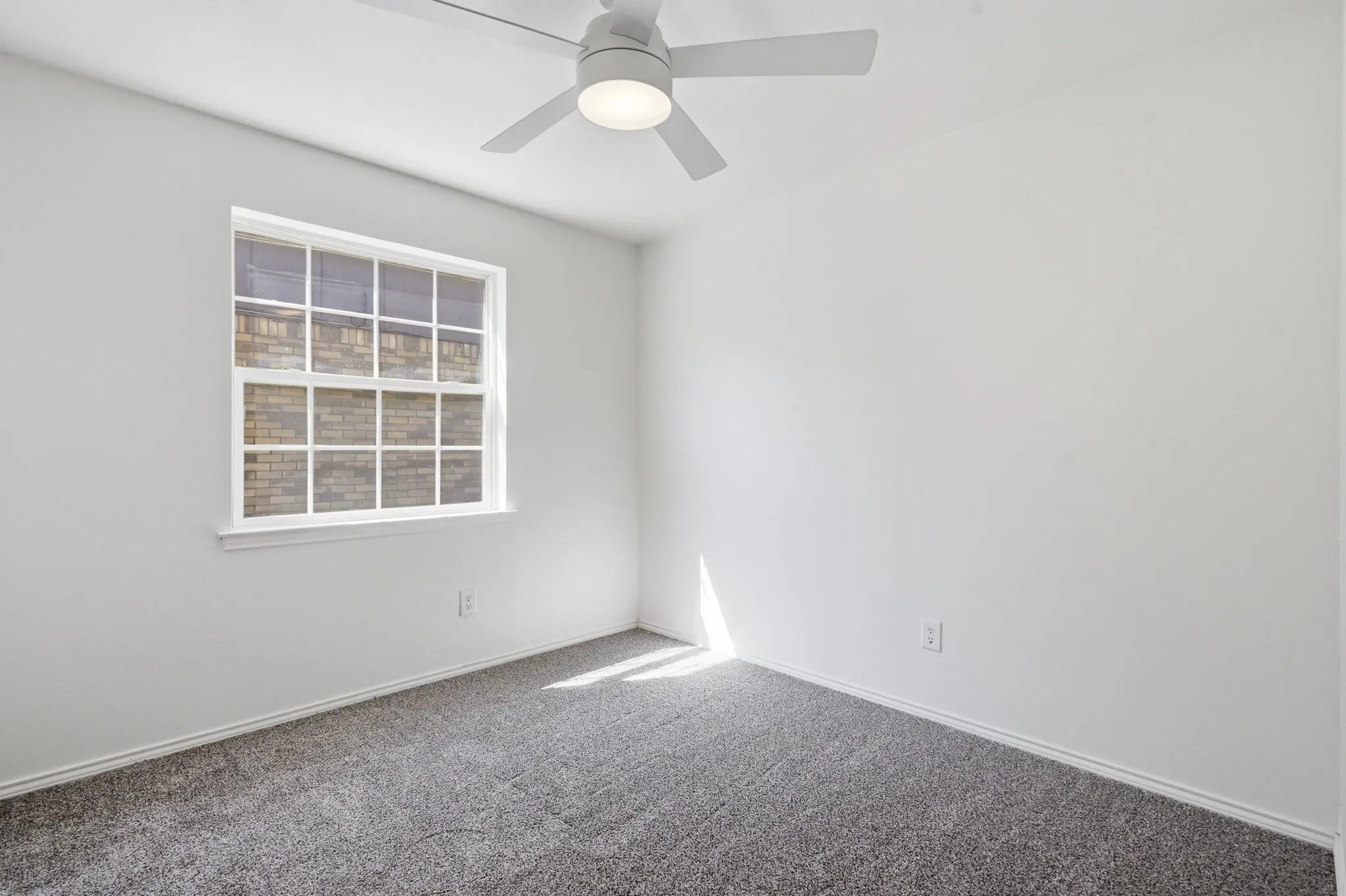 White walls with gray carpet flooring, featuring a window with white sills and grilles, and a ceiling fan with integrated lighting