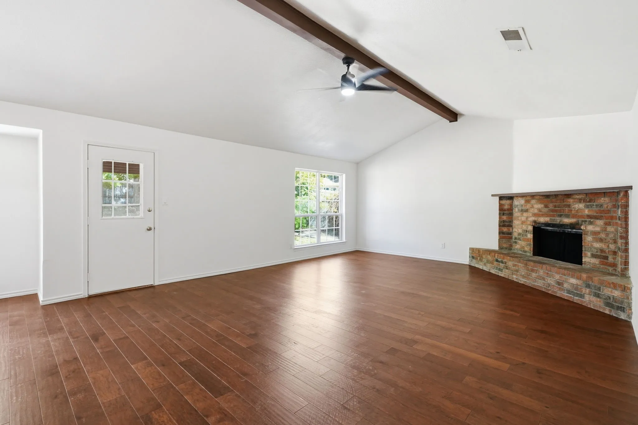 Vaulted ceiling with exposed wood beam, wood-finish flooring, brick fireplace, ceiling fan with light fixture, and multipane windows