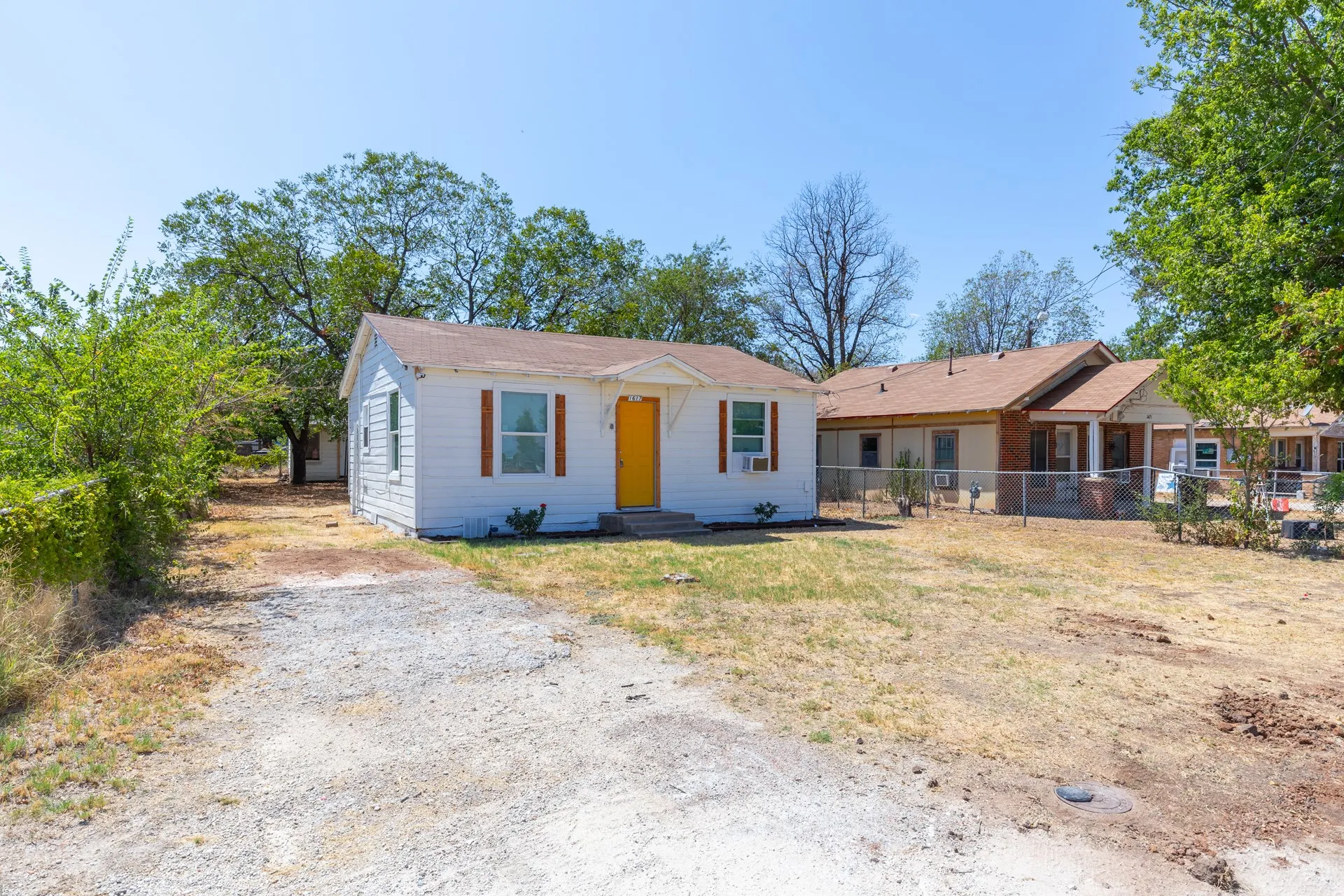 White exterior siding, yellow front door, window shutters, gravel driveway, and mature trees