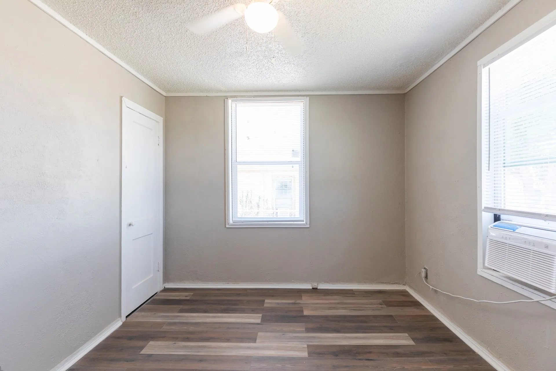 Wood-finish flooring, two windows with blinds, a ceiling fan with integrated lighting, and a white interior door