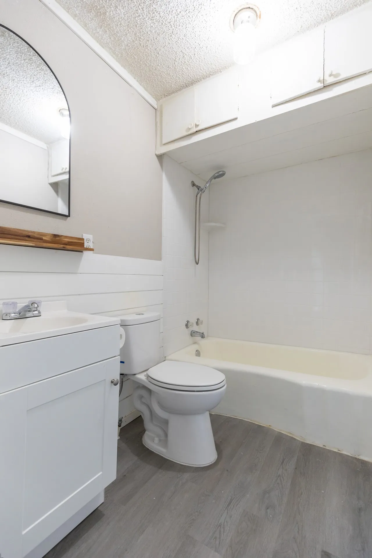White vanity with integrated sink, modern arched mirror, full bathtub with shower fixture, over-tub cabinetry, and wood-finish flooring