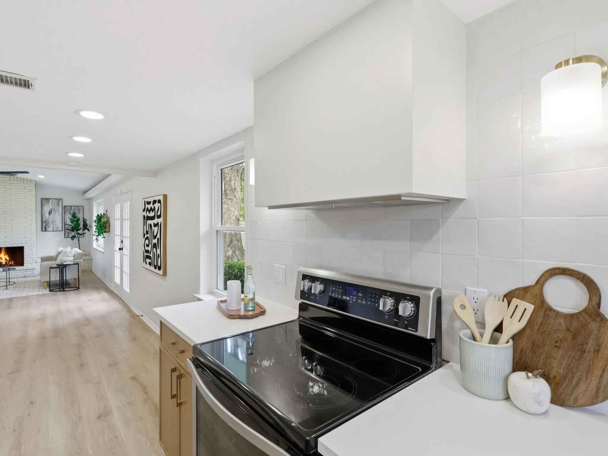 White tile backsplash, stainless steel range with overhead vent, light wood-finish flooring, recessed ceiling lighting, and a white brick fireplace