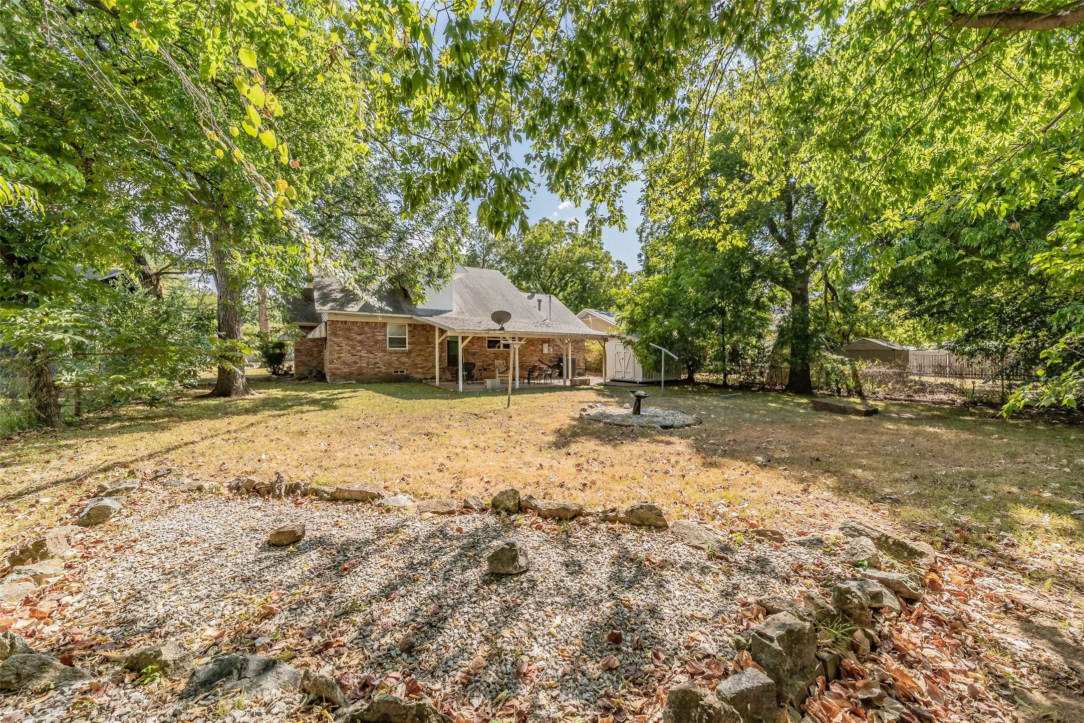 Expansive backyard featuring mature trees, a covered patio, and a dedicated gravel area with rock border