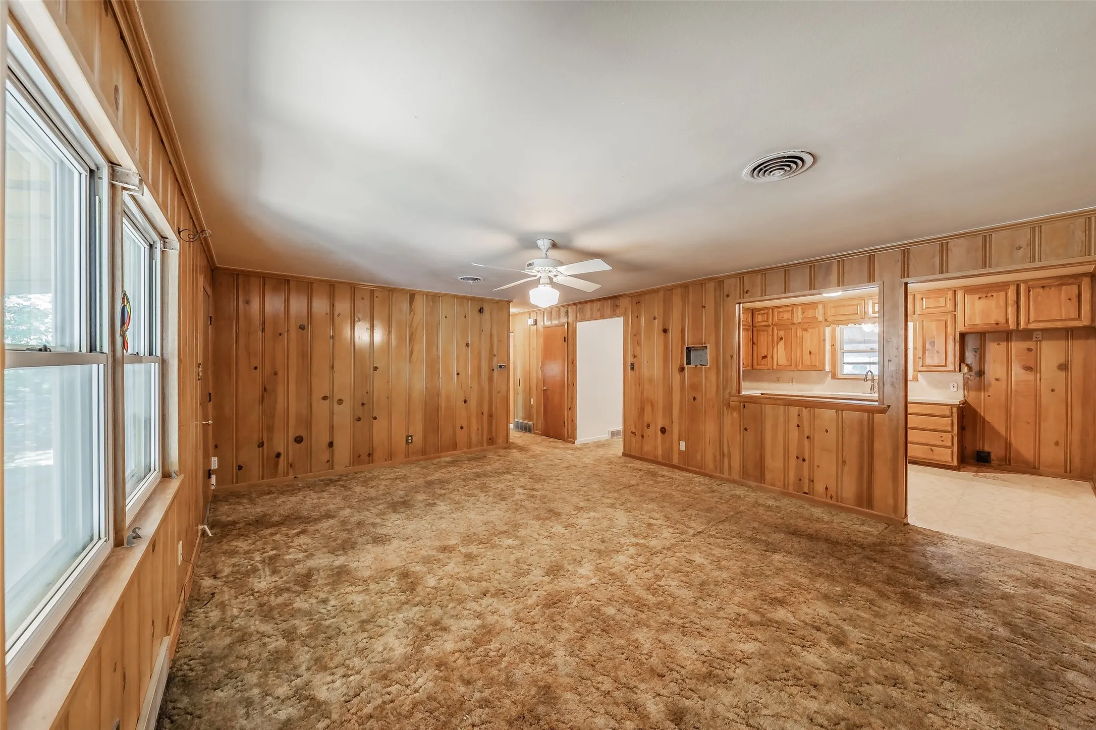 Wood-paneled living space featuring a ceiling fan, multiple windows, and a pass-through opening to a kitchen with wood cabinetry