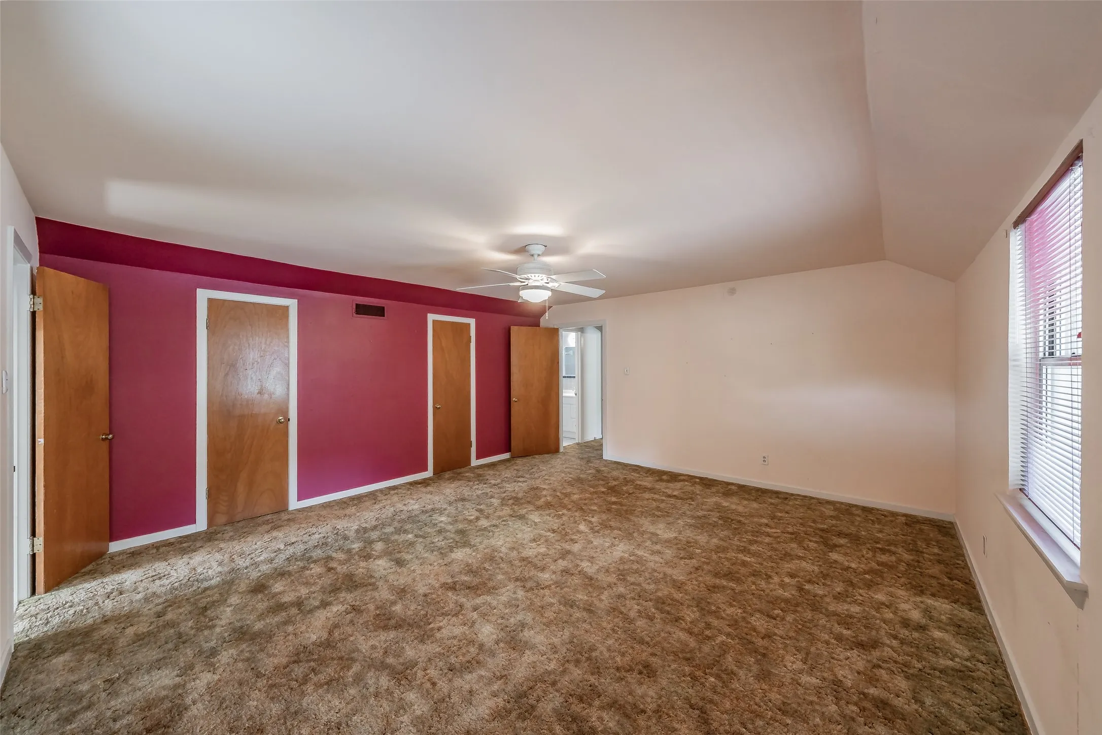 Spacious room featuring sloped ceilings, multiple wood-panel doors, a ceiling fan, and a window with blinds