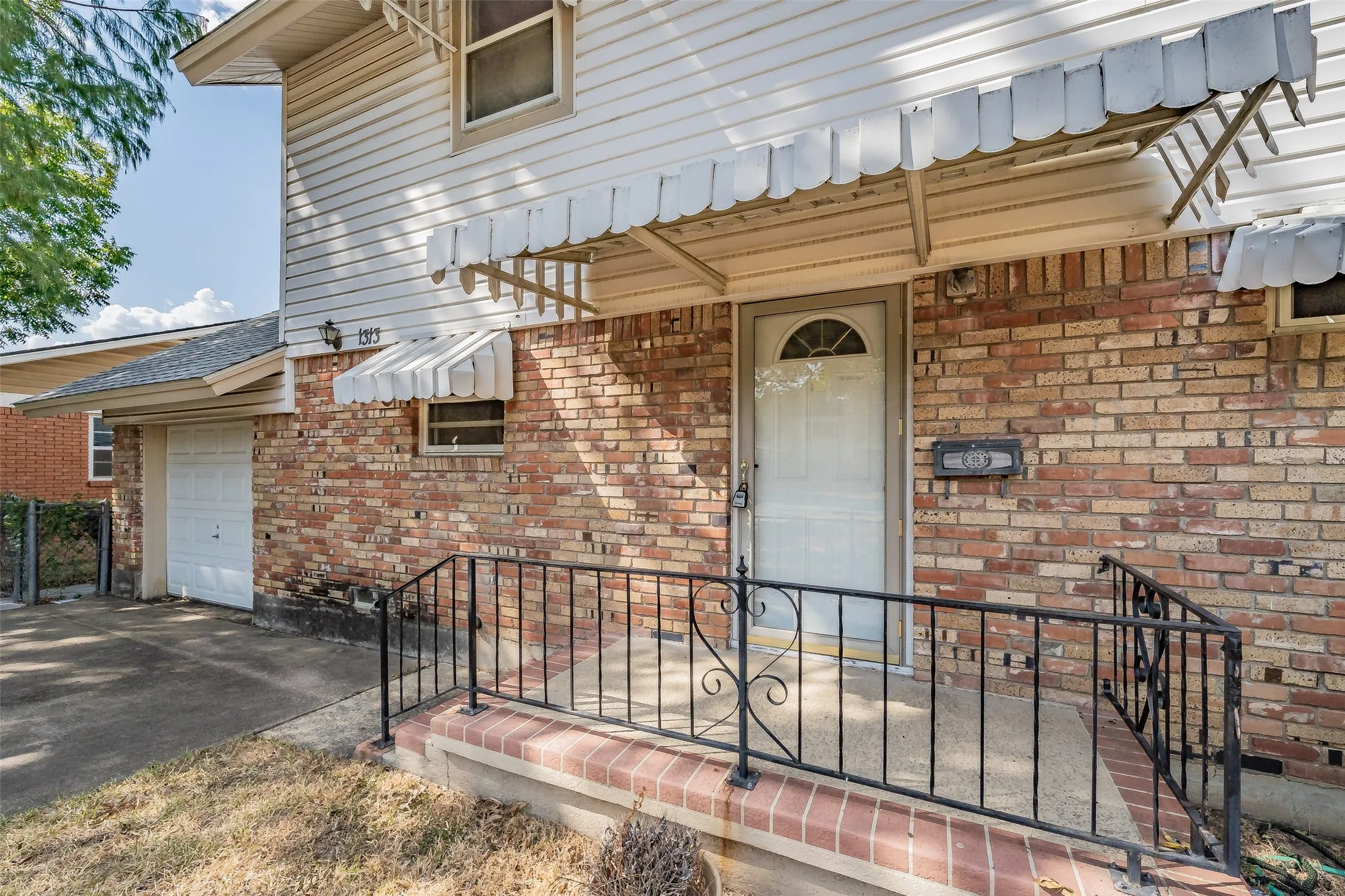 Brick exterior with vinyl siding accents, front entry with storm door, covered porch with wrought iron railing, attached garage with white door, and an exterior awning over the main entry